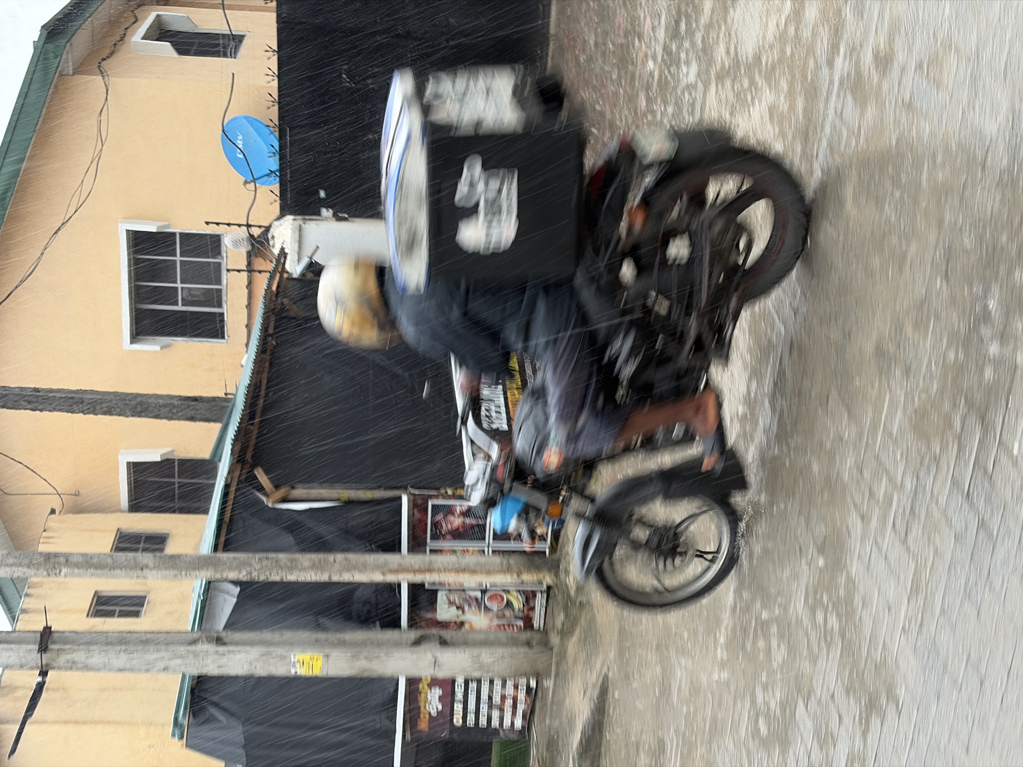 A motorcycle courier, wearing a yellow helmet and a dark jacket, is seen in motion on a wet street during rainfall in Igboefon, Nigeria. The courier is seated on a motorcycle with a large black food delivery box mounted on the rear. The street is paved and appears to be flooded in places. In the background, a yellow building with windows and a green roof is visible, along with a black fence and a blue satellite dish. There are also visible poles and some businesses or stalls with signage. The rain is evident throughout the scene, creating streaks and blurring some details due to motion.