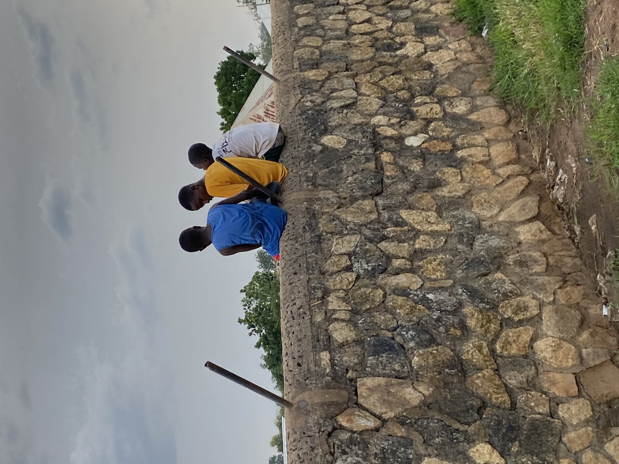 Three young boys are sitting on a rough stone wall, facing away from the camera. The boy on the left wears a blue tank top. The middle boy wears a yellow t-shirt, and the boy on the right wears a white t-shirt with some text and graphics on it. They appear to be holding sticks or similar objects. The wall is made of irregularly shaped stones of varying sizes and colors, mortared together. Two dark metal poles protrude from the wall, one on the left and one on the right, angled slightly upwards. Behind the wall, there are trees, a tent-like structure, and a cloudy sky. The ground in front of the wall is covered with dry dirt and some sparse green grass. The scene is outdoors, suggesting a rural or semi-urban environment in Bukuru, Nigeria.