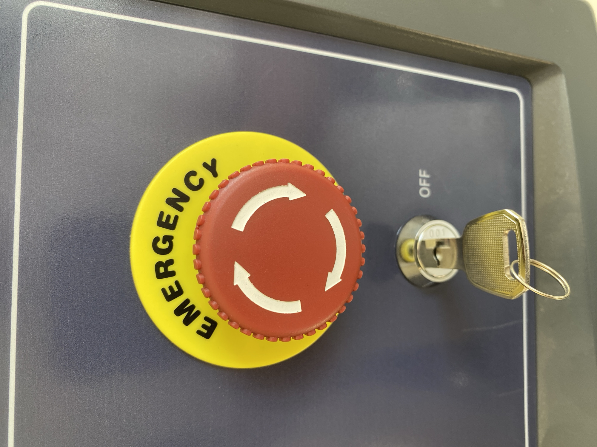 A close-up, slightly angled shot captures a segment of a control panel, featuring an emergency stop button and a key switch. The panel itself is a dark, matte grey or blue-grey color, with a thin white line creating a rectangular border on its surface.
The central subject is a prominent emergency stop button, positioned towards the left. It consists of a textured, red, circular button with a serrated edge, set upon a larger, bright yellow circular base. Three white, curved arrows are visible on the red button, forming a circular pattern, typically indicating a twist-to-release mechanism. The word "EMERGENCY" is printed in black, following the curve of the yellow base, positioned below the red button.
To the right of the emergency stop button, a key switch is visible. A silver key is inserted into the keyhole. The key itself has a metallic brass or gold tone and is connected to a small, simple circular keyring. Above the key switch, the word "OFF" is printed vertically in white text. The number "001" is faintly discernible on the silver cylinder of the key switch mechanism.
The overall lighting is bright and even, suggesting an indoor environment. No people or specific location cues beyond the control panel are visible. The objects imply a safety and control interface for machinery or equipment.