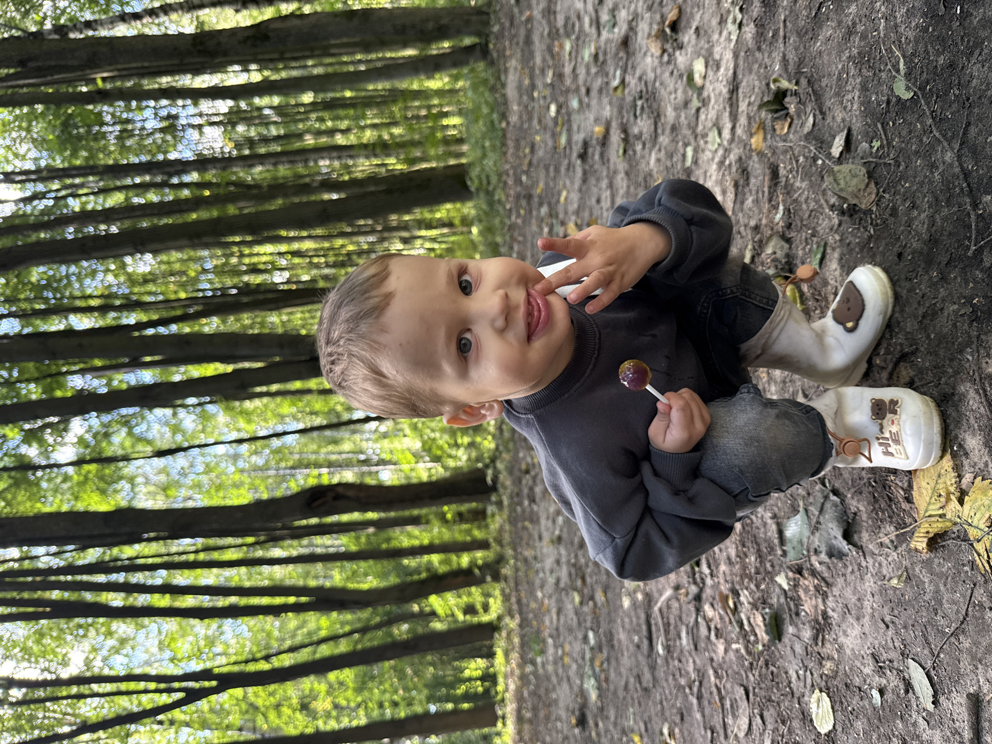 A young boy is crouching in a forest, holding a lollipop. He is wearing a dark grey sweatshirt, black jeans, and white rubber boots. The boy has light brown hair and blue eyes. The forest is filled with tall, thin trees, and the ground is covered in dirt and fallen leaves. The sun is shining through the trees, creating a dappled light effect. The boy appears to be around 3-4 years old. The location is the city of Rusanovo, Russia.