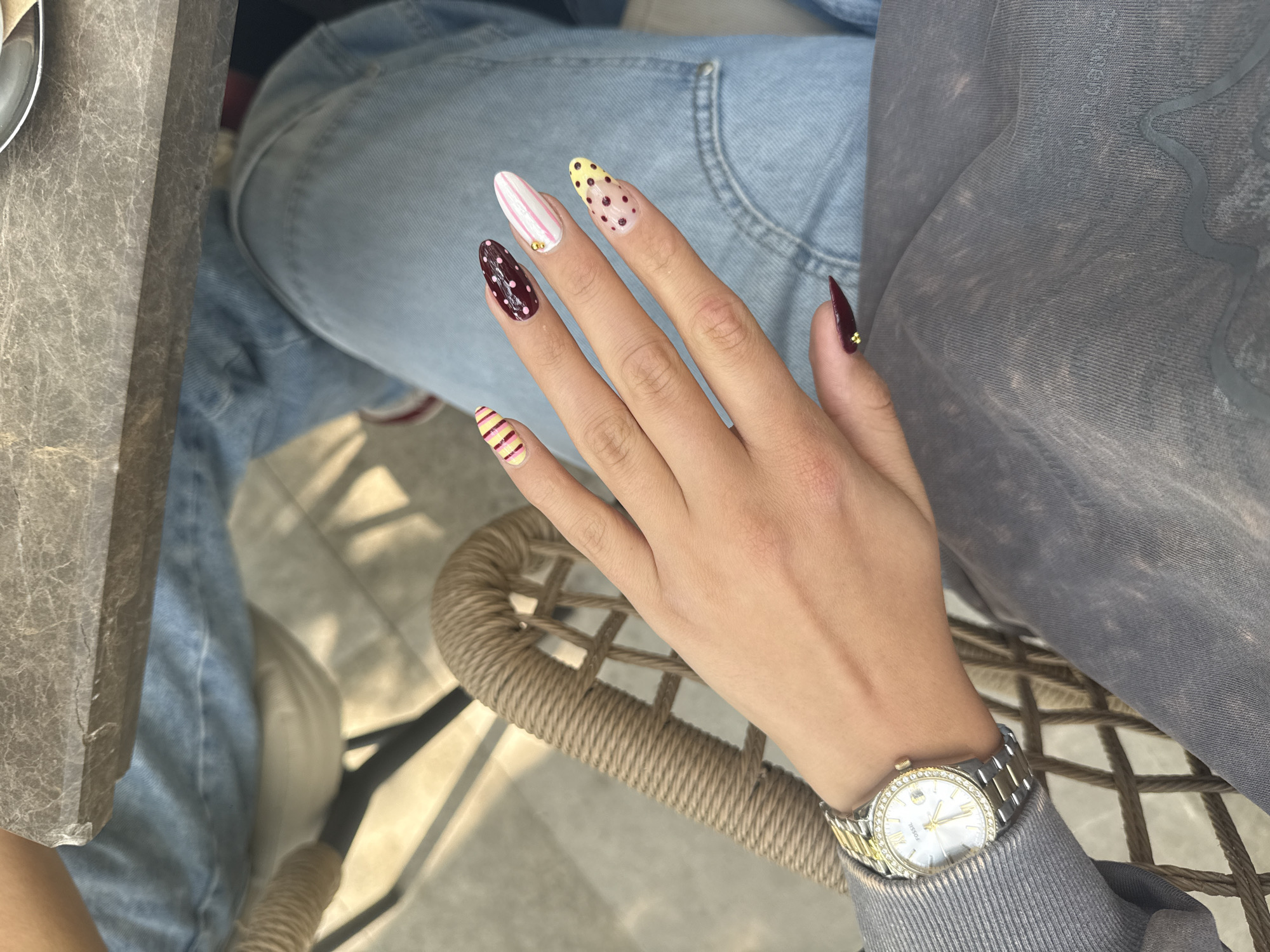 A well-lit, high-angle shot shows a person seated, with their left hand prominently displayed. The hand, adorned with a silver and gold watch on the wrist, rests against a wicker chair. The person wears light blue denim jeans and a gray sweatshirt with a faded pattern. The fingernails are each painted with a different pattern in shades of red, yellow, white, and brown. The background consists of a gray tabletop on the left and the woven texture of the chair's seat. The image provides no specific details that confirm the location being Plovdiv, Bulgaria.