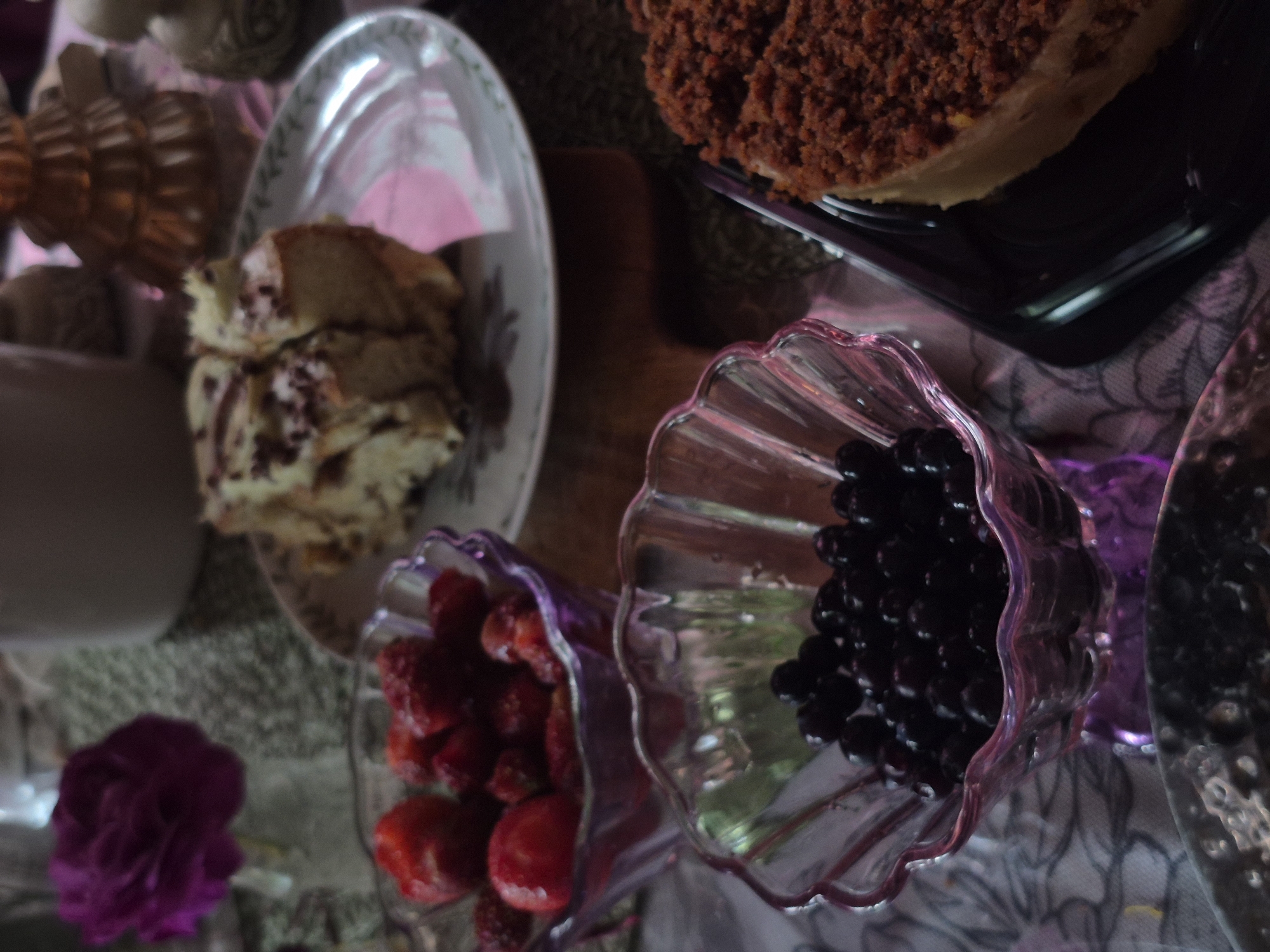 The image depicts a close-up, overhead view of a table setting featuring desserts and fruit. In the foreground, a clear glass bowl filled with dark blueberries is visible, set against a floral patterned tablecloth. To the left, another clear glass bowl holds bright red strawberries. Behind these bowls, a slice of cake with chocolate frosting sits on a patterned plate. To the right, a slice of cake with a crumb topping is presented on a black rectangular platter. A single purple rose in a vase is situated in the upper left corner of the frame. Decorative elements, including what appears to be a golden Christmas tree-shaped candle holder, are also present in the background. The lighting is dim, creating a moody atmosphere. The scene is set indoors. The specific location in Астра-1 Днт, Russia is not discernible from the image itself.