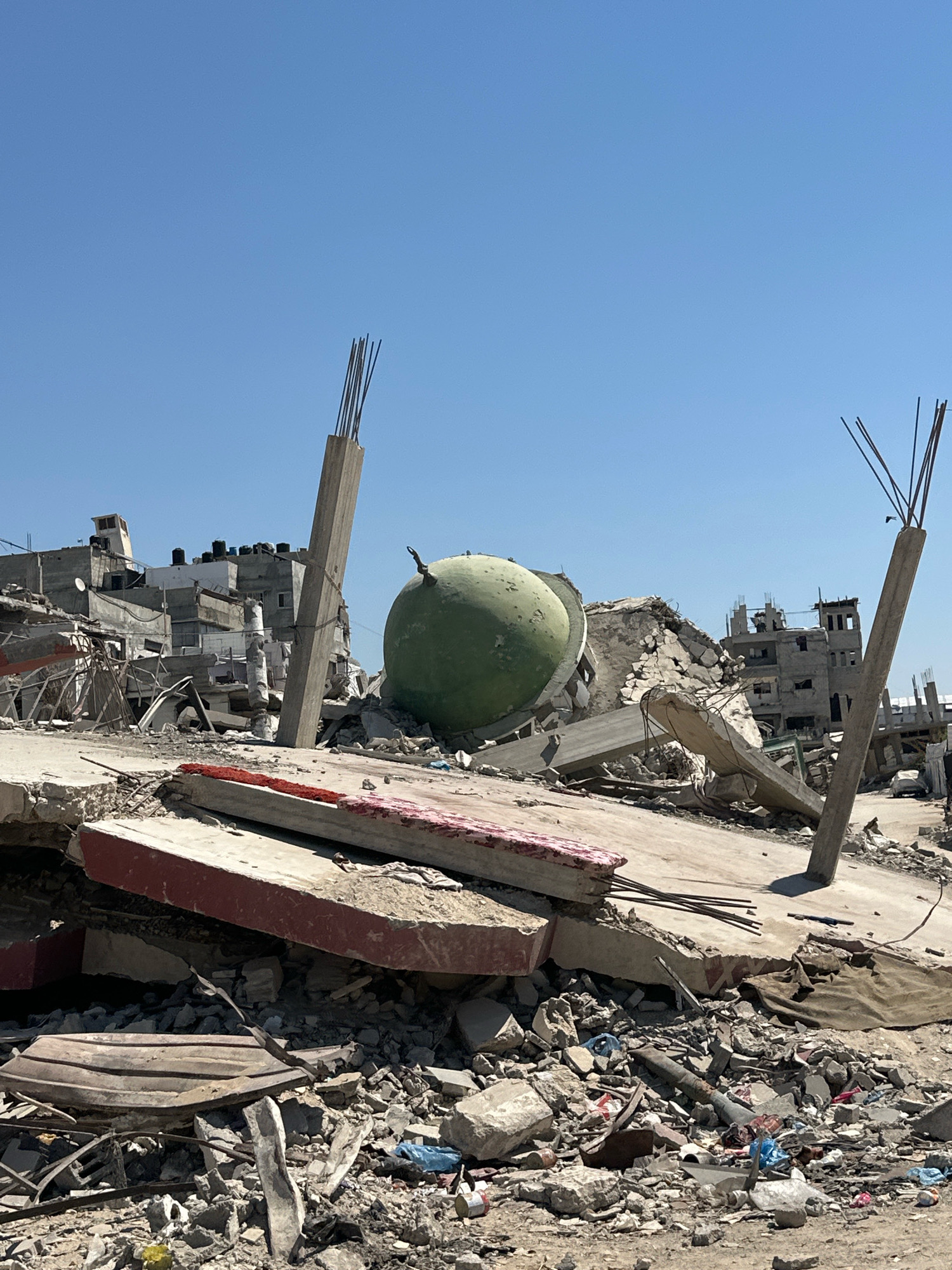 This image depicts the aftermath of destruction in the city of Gaza, Palestinian Territories. The scene is dominated by the remnants of collapsed buildings, with concrete debris, twisted rebar, and rubble covering the ground. In the center of the frame, a large, green, dome-shaped structure is visible, partially embedded in the rubble. Several tall, concrete pillars, some with exposed rebar, stand precariously amidst the destruction, suggesting that these were part of larger structures that have since fallen. In the background, more damaged buildings are visible, some appearing to be multi-story structures. The sky above is clear and blue. The overall impression is one of widespread destruction and devastation within an urban environment. There are no people visible in the image.