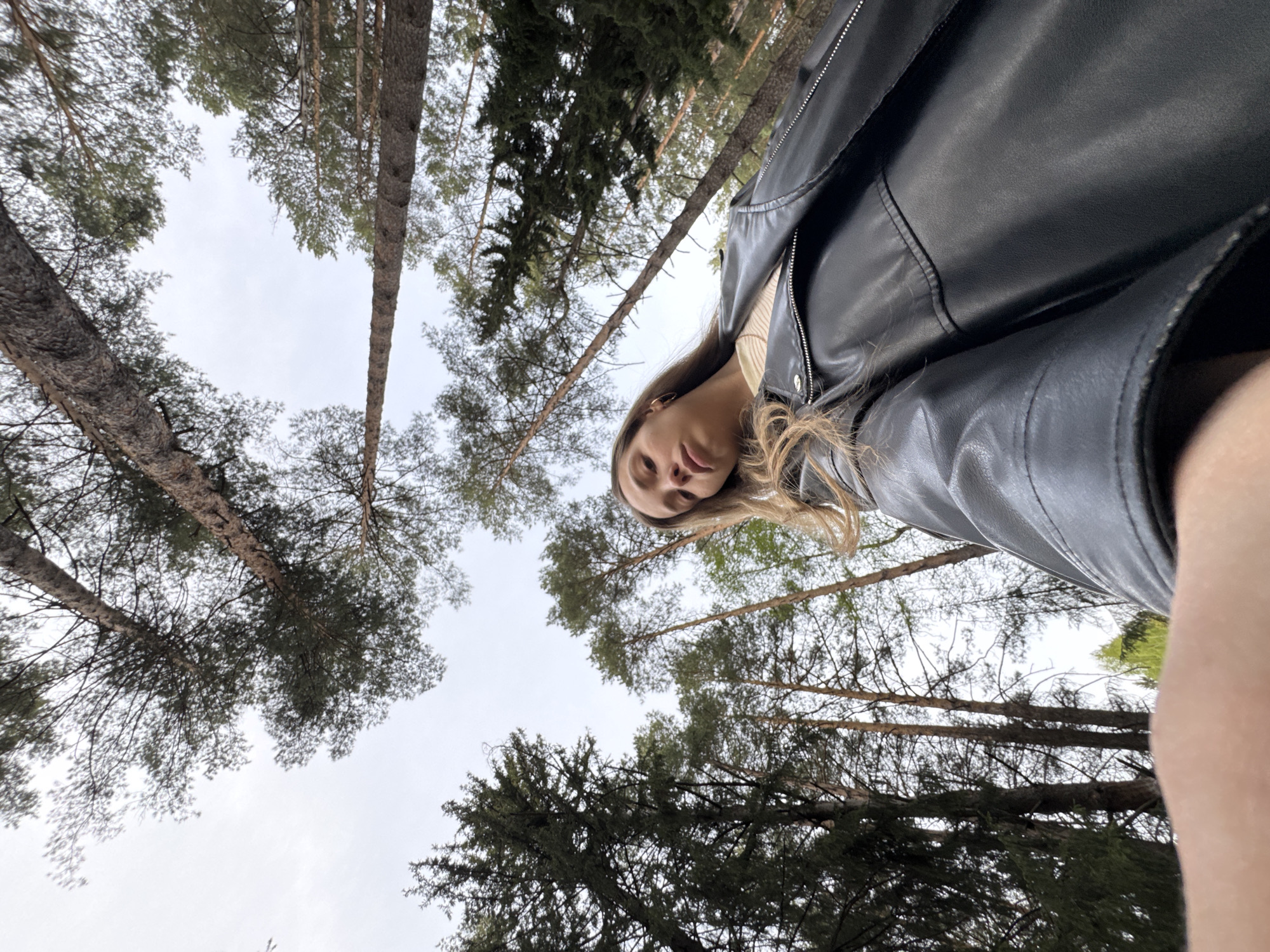 A low-angle shot looking up at a person in a black leather jacket surrounded by tall trees. The location appears to be a forest. The sky is overcast. The person's face is visible, with blonde hair. Based on location of the image this is likely located in Прикамский, Russia.