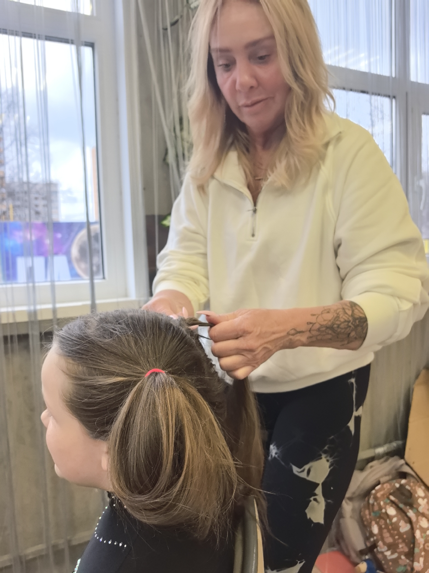 A middle-aged woman with blonde hair is braiding the hair of a young girl inside a room. The girl has brown hair tied back with a red elastic band. The woman is wearing a white zippered sweatshirt and black patterned leggings. The background includes a window with sheer curtains, a view of buildings (possibly residential) outside, and other indistinct items suggesting a domestic or salon setting. The location is Izhevsk, Russia.