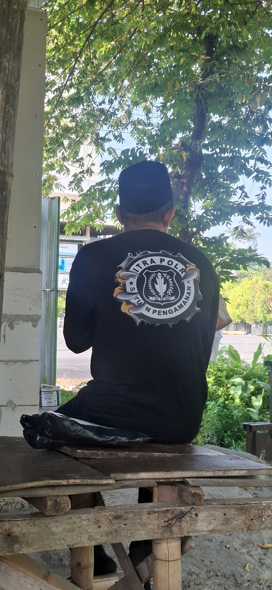 A full-shot image shows a man from behind, seated on a wooden structure outside, under the shade of a tree with green leaves. He is wearing a black long-sleeved shirt with a circular emblem on the back and a black peci hat. The emblem features intricate designs and text, partially obscured by the man's posture. A black plastic bag rests on the wooden surface in front of him. The background reveals a street with sparse greenery, a building with white brick walls on the left, and a blue sky. The man appears to be facing a street, with hints of residential or commercial buildings visible further away. The scene is outdoors, likely in a semi-rural or suburban area of Gajahan, Indonesia, given the architectural style of the nearby building and the general appearance of the surroundings. The lighting suggests it is daytime, with sunlight filtering through the leaves of the tree.