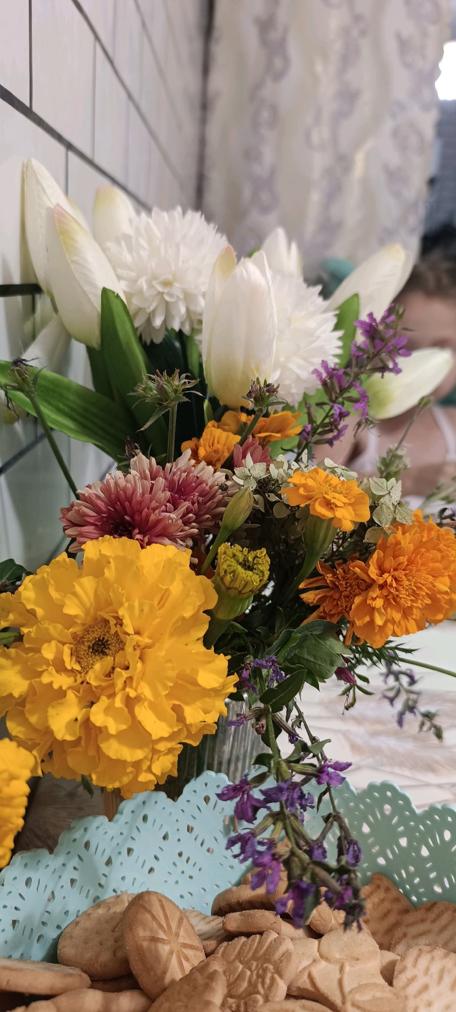 The image shows a close-up of a bouquet of flowers in a clear glass vase, placed on a table. The bouquet features a large yellow marigold prominently in the foreground, with smaller orange marigolds and pinkish-red dahlias, as well as white tulips and smaller purple flowers. The background is blurred, showing a white tiled wall and a light-colored curtain. Below the vase, a light blue decorative doily is visible, underneath which are several cookies. The lighting suggests an indoor setting, possibly a home or cafe. The scene does not depict anything specific to the city of Chaikovsky, Russia.