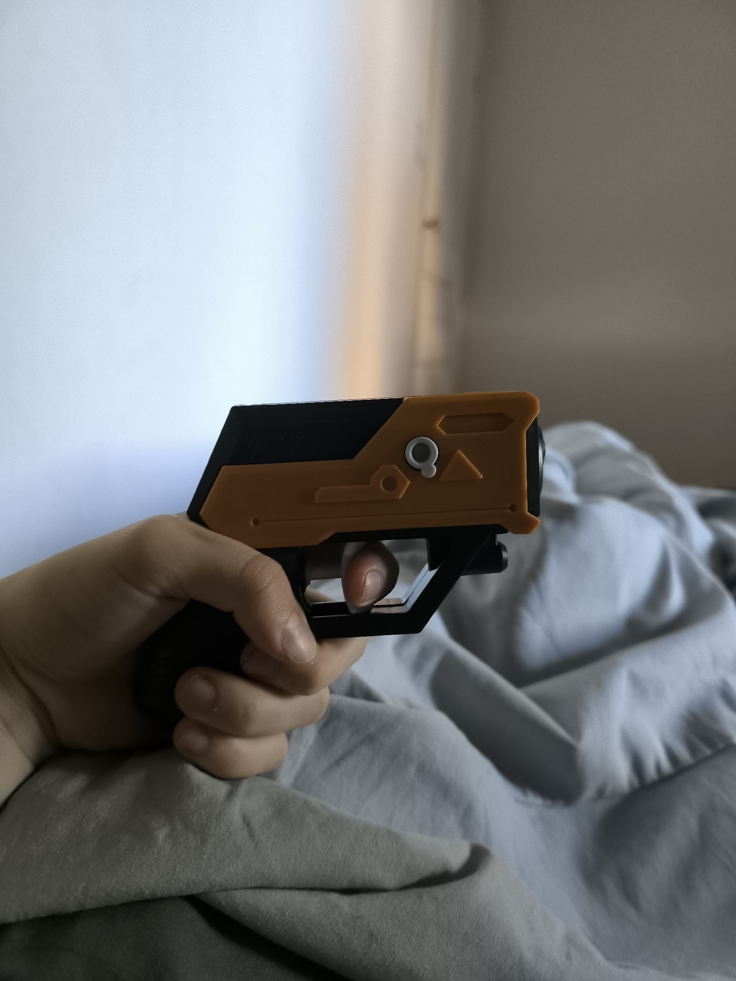 A hand holds a black and orange plastic toy gun on top of a gray bed sheet. The background consists of a white wall partially illuminated by sunlight. The location is not discernible from the image.