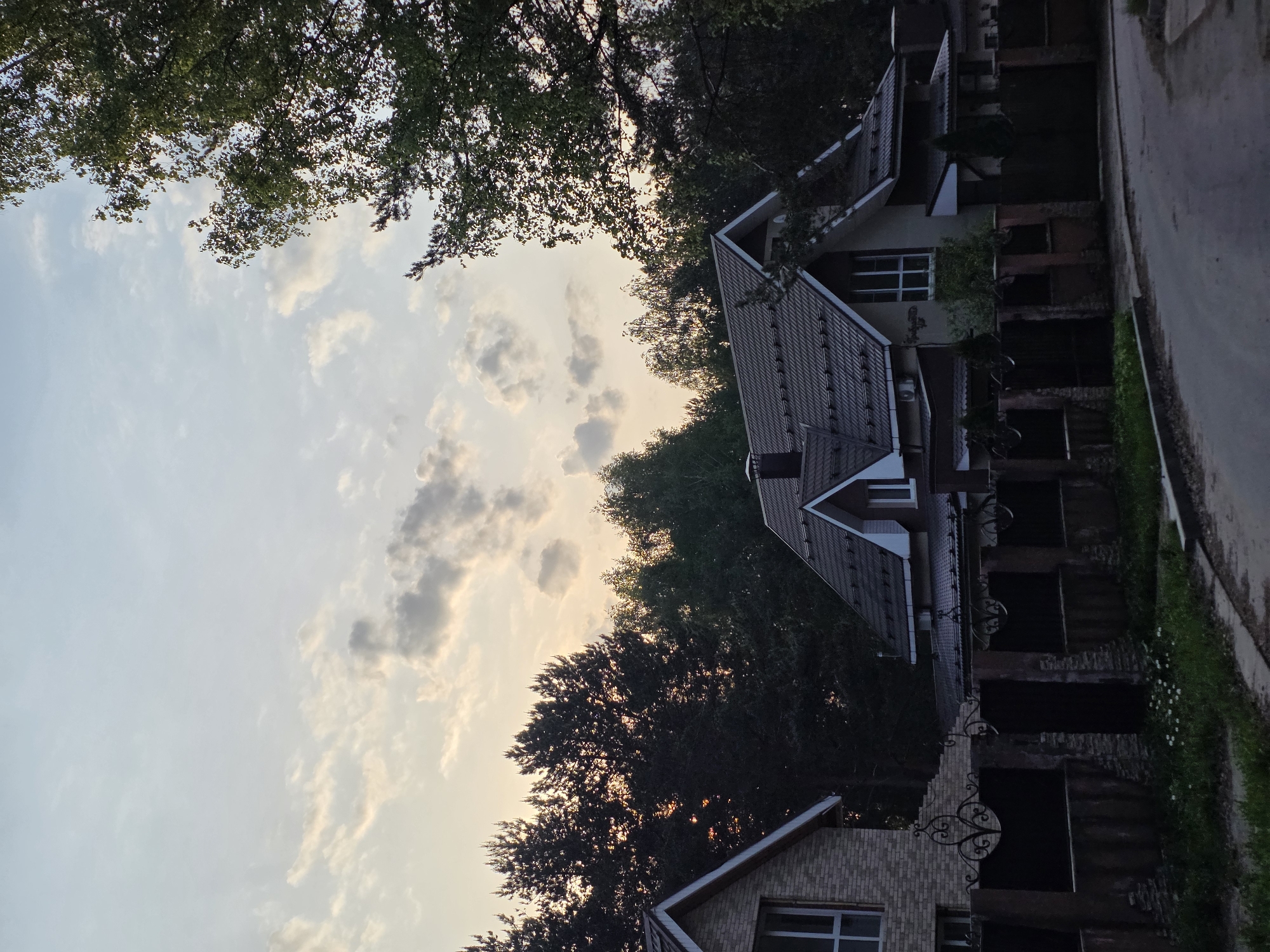 The image presents a view of a residential area in what is likely a suburban or semi-rural part of the city of Chaikovsky, Russia, based on the architecture and vegetation typical of that region.
The perspective is looking up at the sky, framed by trees and the rooftops of several houses. The sky is a pale blue color with scattered clouds, possibly at dusk or dawn, judging by the soft lighting.
The houses exhibit a mix of architectural styles, including gabled roofs, brick facades, and visible windows. There is a wrought-iron fence or decorative element along what appears to be a walkway or patio area of one of the houses. The image is rotated. A road or street is visible alongside the houses.