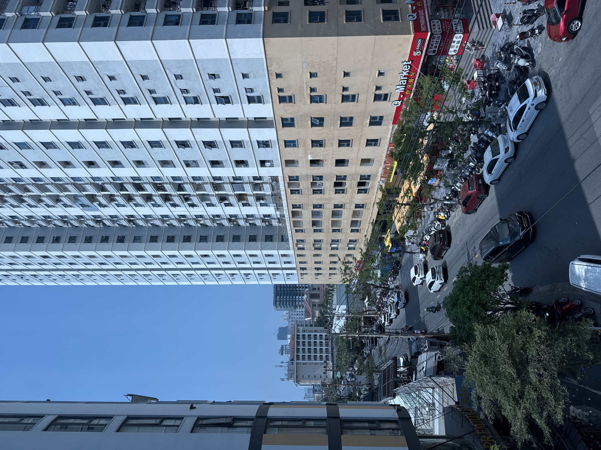 This is an eye-level shot of a street in Đà Nẵng, Vietnam. The sky is a clear, pale blue. The street is lined with tall buildings, one a high-rise apartment complex with rows of identical windows. Next to it is another building with many windows. The street is filled with motorbikes and cars parked along the side. A few pedestrians are visible. A "Q-Market" sign is visible on one of the buildings.