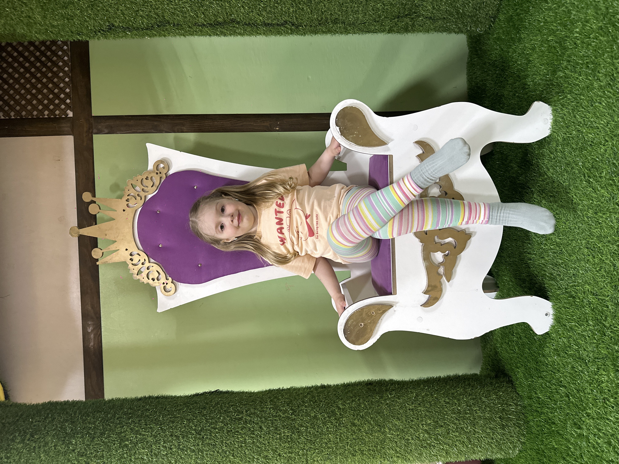 A young girl is sitting in a white and purple princess throne chair, located in what appears to be an indoor play area. The area has artificial grass on the floor and parts of the wall. The wall is also partly green. The location is Glazov, Russia. The girl has light hair, a light orange shirt with text, and striped pastel-colored leggings. She is wearing socks and smiling. The throne has a golden crown decoration on its left side and cut-out patterns on the lower part of the chair. The background includes a wooden and light-colored wall section above the green portion.
