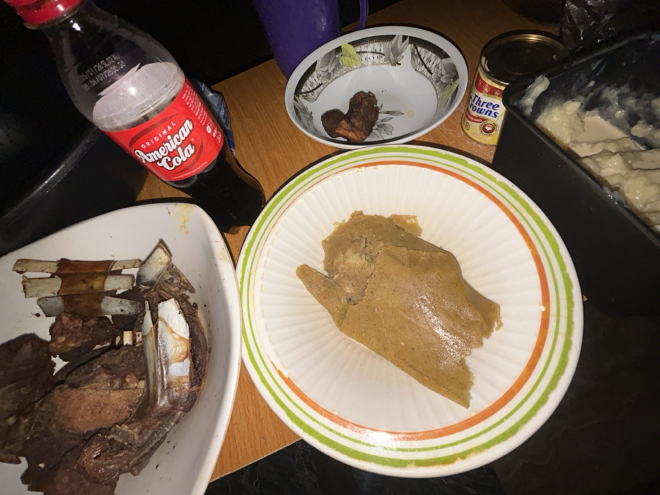 The scene depicts a tabletop arrangement of food and drink in Debasco, Nigeria. In the upper left, a white, irregularly shaped serving dish contains a substantial quantity of dark, cooked meat, including ribs with visible bones. Below this, a white bowl with green and orange concentric stripes on its rim holds a large, light brown, paste-like food item, consistent with a traditional Nigerian meal component. To the right, a smaller white bowl with a floral/geometric pattern contains a few dark, small pieces of cooked food. A plastic bottle of "American Cola" with a red cap is partially visible, positioned diagonally on the table, indicating it has been consumed. Further right, a yellow can labeled "Three Crowns" is present, alongside a dark container holding a pale, textured substance. The table surface is light brown wood, and the background is dark, suggesting an indoor setting with ambient lighting. No individuals are present in the image.