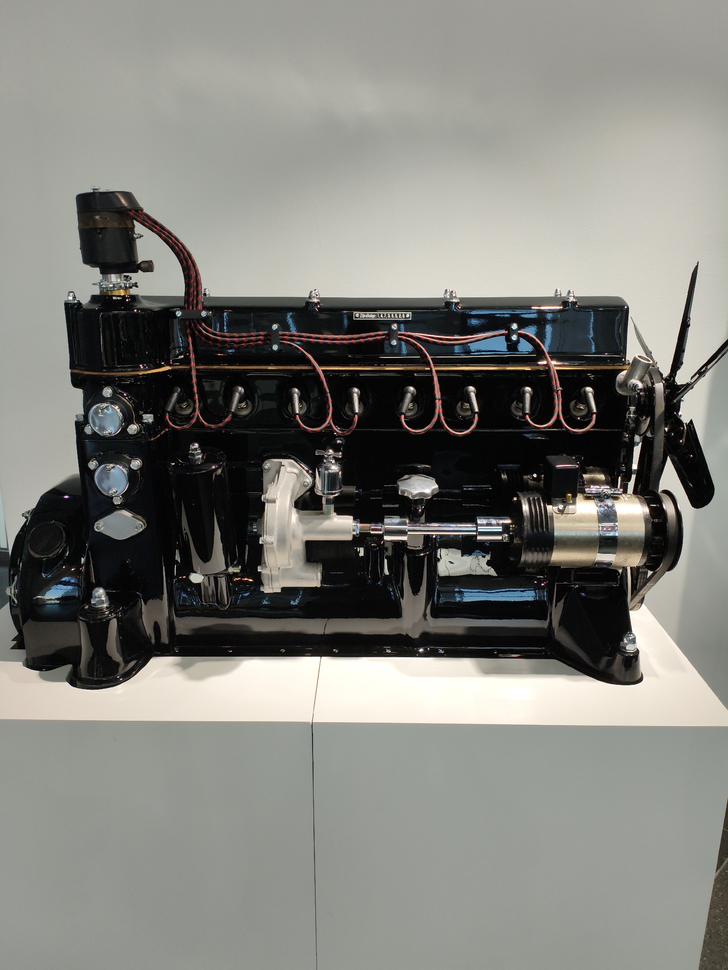 An antique, black, straight-six internal combustion engine is displayed on a white pedestal in a museum exhibit. The engine is polished and appears to be in excellent condition, with visible spark plug wires, a distributor cap, a fan, and other components. A small label on the engine reads "Zündfolge: 1.4.7.3.6.5.2.8". The background is a plain, light gray wall. This exhibit is located in Ingolstadt, Germany.