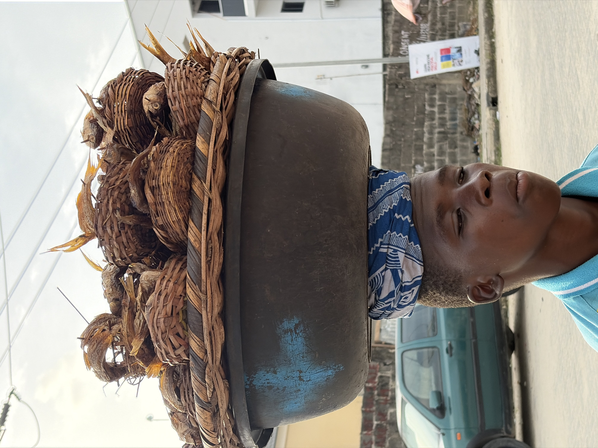 A young person, likely male, with dark skin and short hair, is carrying a significant load on his head in what appears to be a street scene in Igboefon, Nigeria. He wears a blue and white patterned headwrap underneath a large, dark, worn basin, which serves as the base for the load. Piled on top of the basin are numerous smaller, intricately woven baskets made of dried brown plant fibers. Each basket contains several dried fish, clearly identifiable by their shape, scales, and fins, indicating they are goods for sale or transport. The person's gaze is directed slightly upward and forward, with a calm, focused expression. He wears a light blue polo shirt with white trim.
The setting is outdoors during the daytime, likely under a bright, possibly overcast sky, visible in the upper left. The background features a paved road or sidewalk. To the right, a building with unplastered concrete blocks is visible, against which a white sign leans. Partial text on the sign includes "ONSENT" and "DIGITAL," along with four colored squares (red, yellow, blue, white). A light teal-green van or small bus is partially visible behind the person, parked on the street. Some debris is present on the ground near the building. The image captures a common practice of head-loading goods in many African communities.