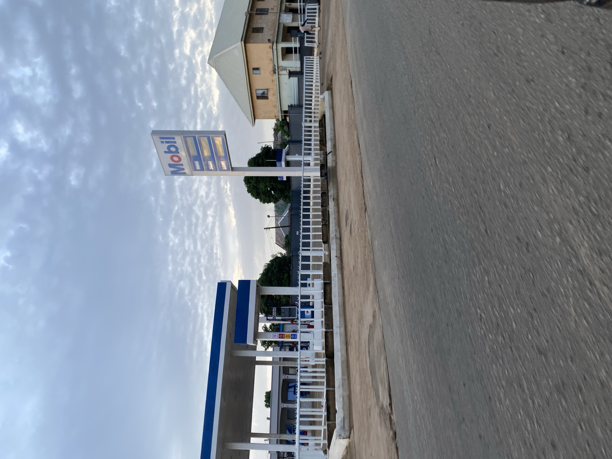 The image depicts a Mobil gas station in Jos, Nigeria, as indicated by the Mobil sign and the architectural style of the buildings, which is consistent with structures found in West Africa. The gas station features a covered forecourt with blue and white accents, and several fuel pumps are visible. A white railing encloses the gas station premises. To the right of the gas station is a multi-story building with a light-colored facade and dark-rimmed windows. A person in light-colored clothing is visible walking near the entrance of this building. The sky is overcast with scattered clouds. The foreground shows a paved road. The scene appears to be captured during daylight hours, possibly early morning or late afternoon, given the diffused lighting and cloudy sky.