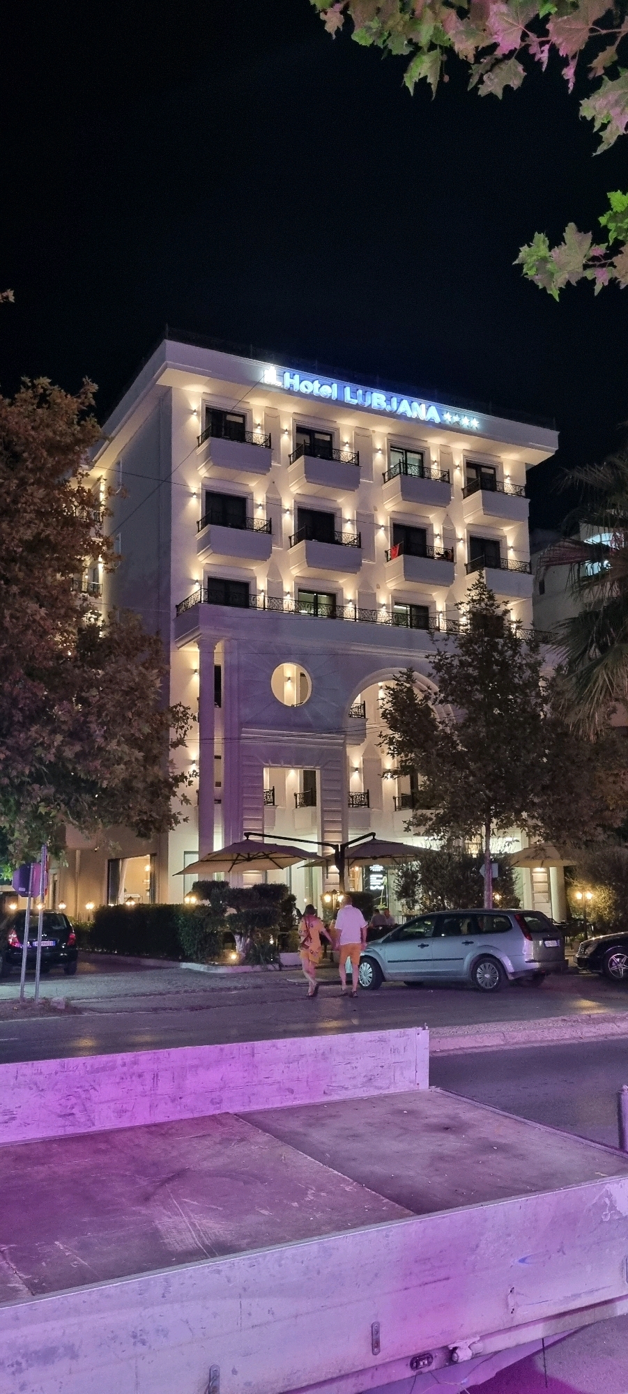 The image shows the exterior of Hotel LUBJANA at night, located in Bashkia Durrës, Albania. The hotel is a multi-story building with a white facade, illuminated by numerous lights. The hotel's name is visible in blue neon letters on the top of the building, along with three stars. Several balconies with black railings are visible on the upper floors. In front of the hotel, there are two large umbrellas, suggesting an outdoor seating area.
In the foreground, a light-colored car is parked on the street, and a silver station wagon is also visible. Two individuals, a man and a woman, are walking away from the camera on the sidewalk. The woman is wearing a yellow dress, and the man is wearing a white shirt and orange shorts. There are trees with foliage surrounding the hotel and the sidewalk. The street in front of the hotel is wet, reflecting the lights, and a concrete structure with purple lighting is in the extreme foreground. The sky is dark, indicating nighttime.