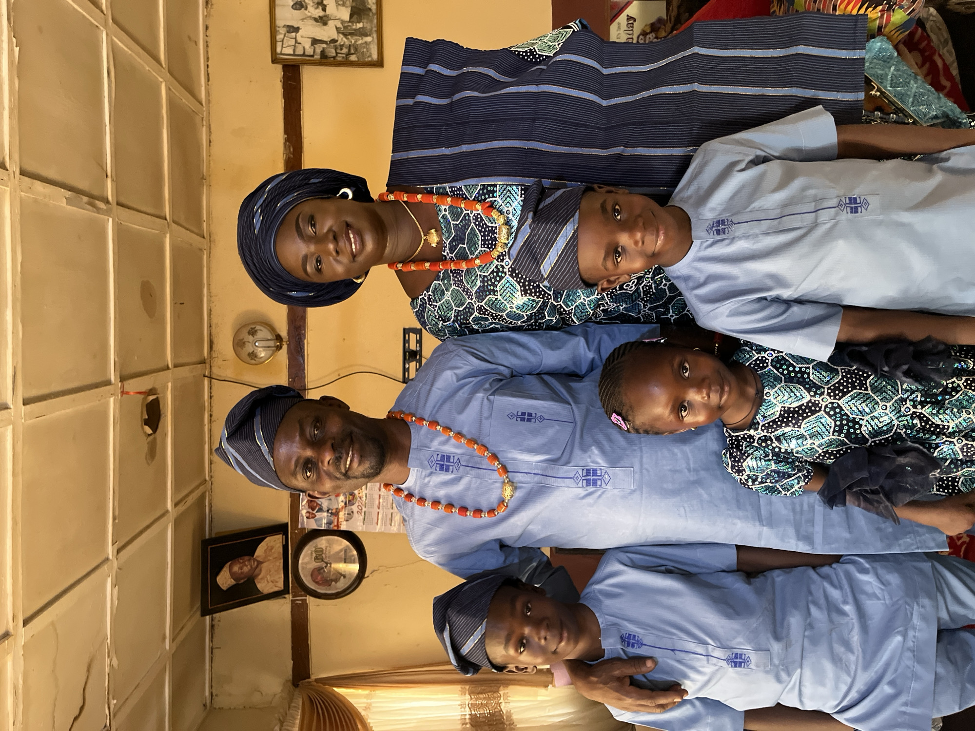 In Rafin Bauna, Nigeria, a family of five poses for a photo indoors. A man and a woman stand side-by-side, each wearing traditional attire including headwear and coral bead necklaces. The man wears a light blue tunic, while the woman wears a patterned top and a dark blue headwrap. Three children are positioned in front of them. Two boys wear light blue tunics and matching headwear, while a girl wears a patterned top. The background includes framed photos on the wall, a calendar, and a partially visible window. The room has off-white walls with noticeable imperfections.
