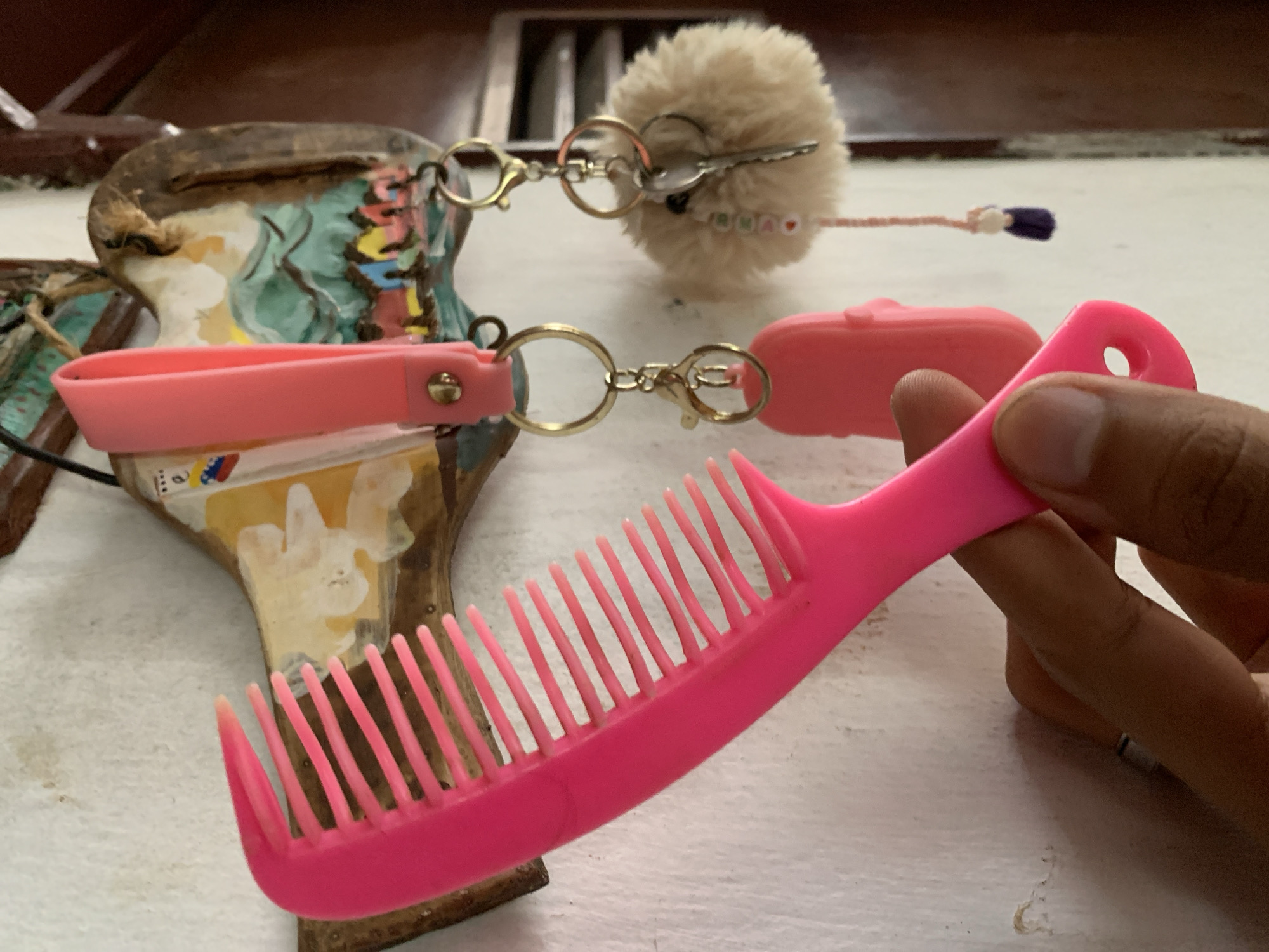 The image shows a pink comb held by a hand. Behind the comb, there are various objects including a decorative carved object, pink loop and case attached to a metal ring, and a fluffy keychain. The background is a blurred surface with wood trim. It is impossible to determine the location in Santa Ana, Venezuela from the image.