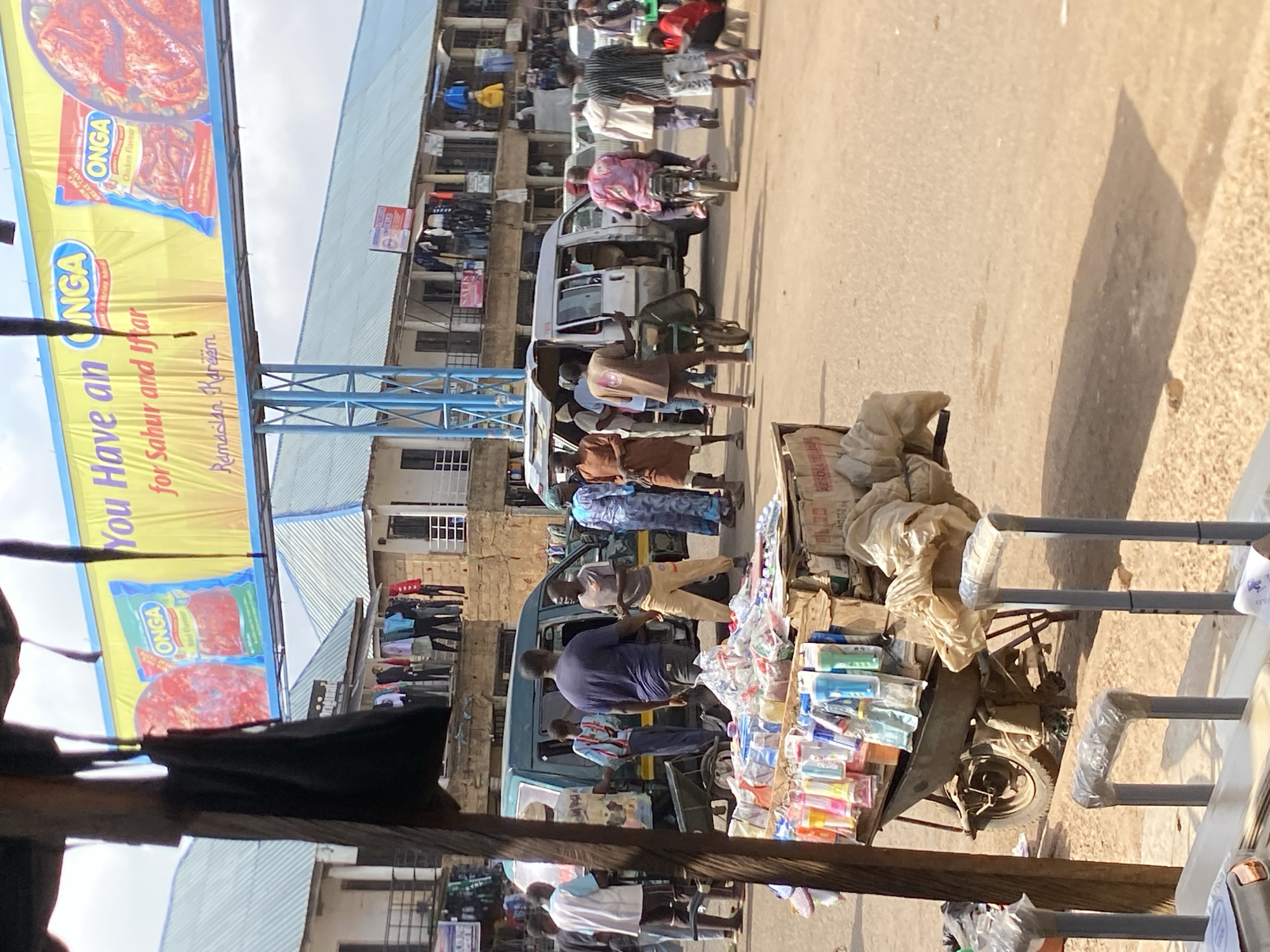 The image depicts a busy street scene in what appears to be Bukuru, Nigeria. People are walking along the street, near buildings and shops. A white van is parked in the middle of the road, with people standing and walking around it. A hand-pushed cart laden with goods is parked on the side, partially obscured by what seems to be a railing. Overhead, a large advertisement for "ONGA" food seasoning, advertising "for Sahur and Iftar Ramadan Kareem" is prominently displayed. Clothes are hanging from some buildings.