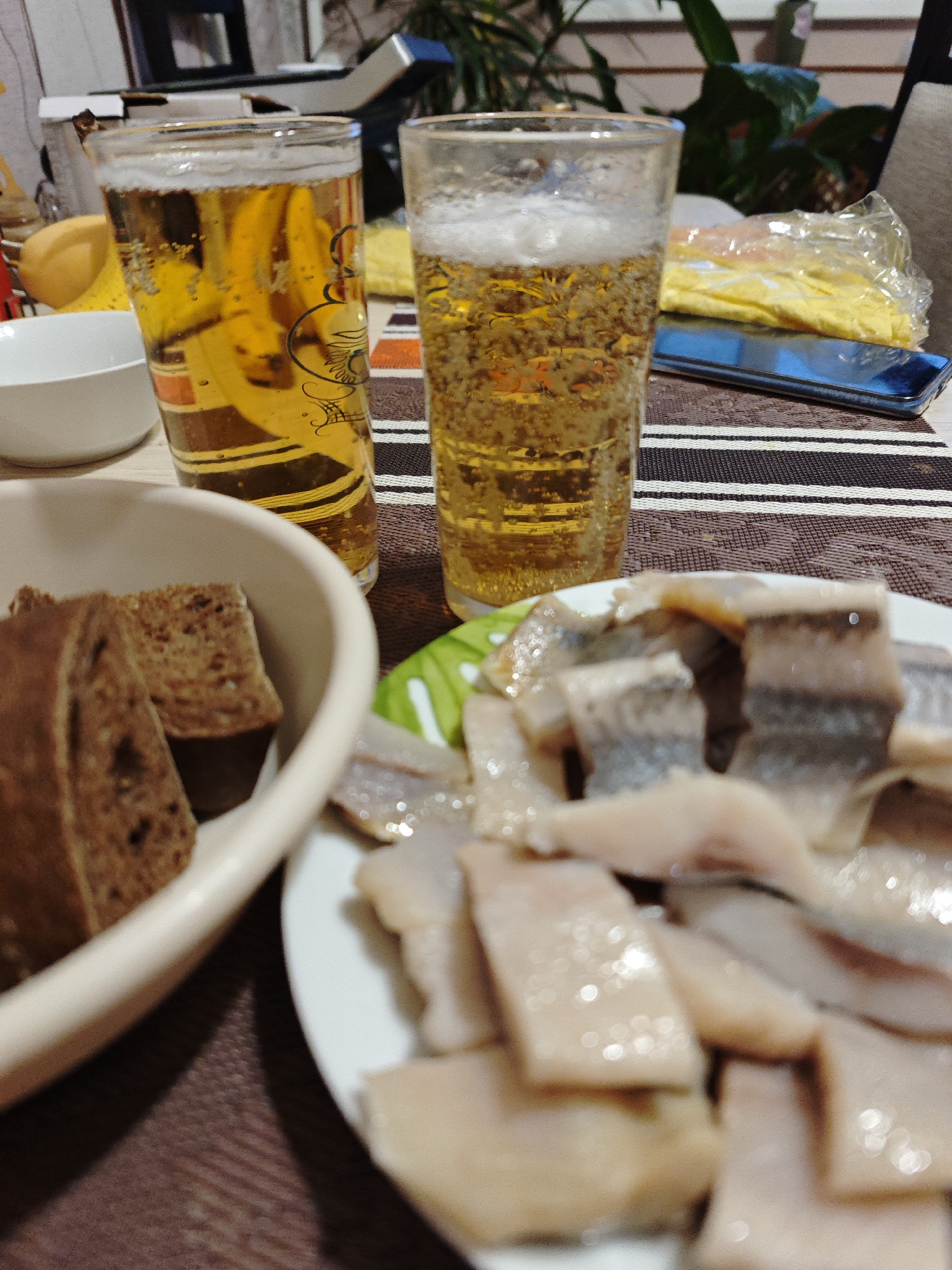 A table setting with two tall glasses of beer, a bowl of dark bread, and a plate of sliced herring. A blue smartphone is visible in the background on a brown and white striped tablecloth. The location is presumed to be Марковский, Russia.