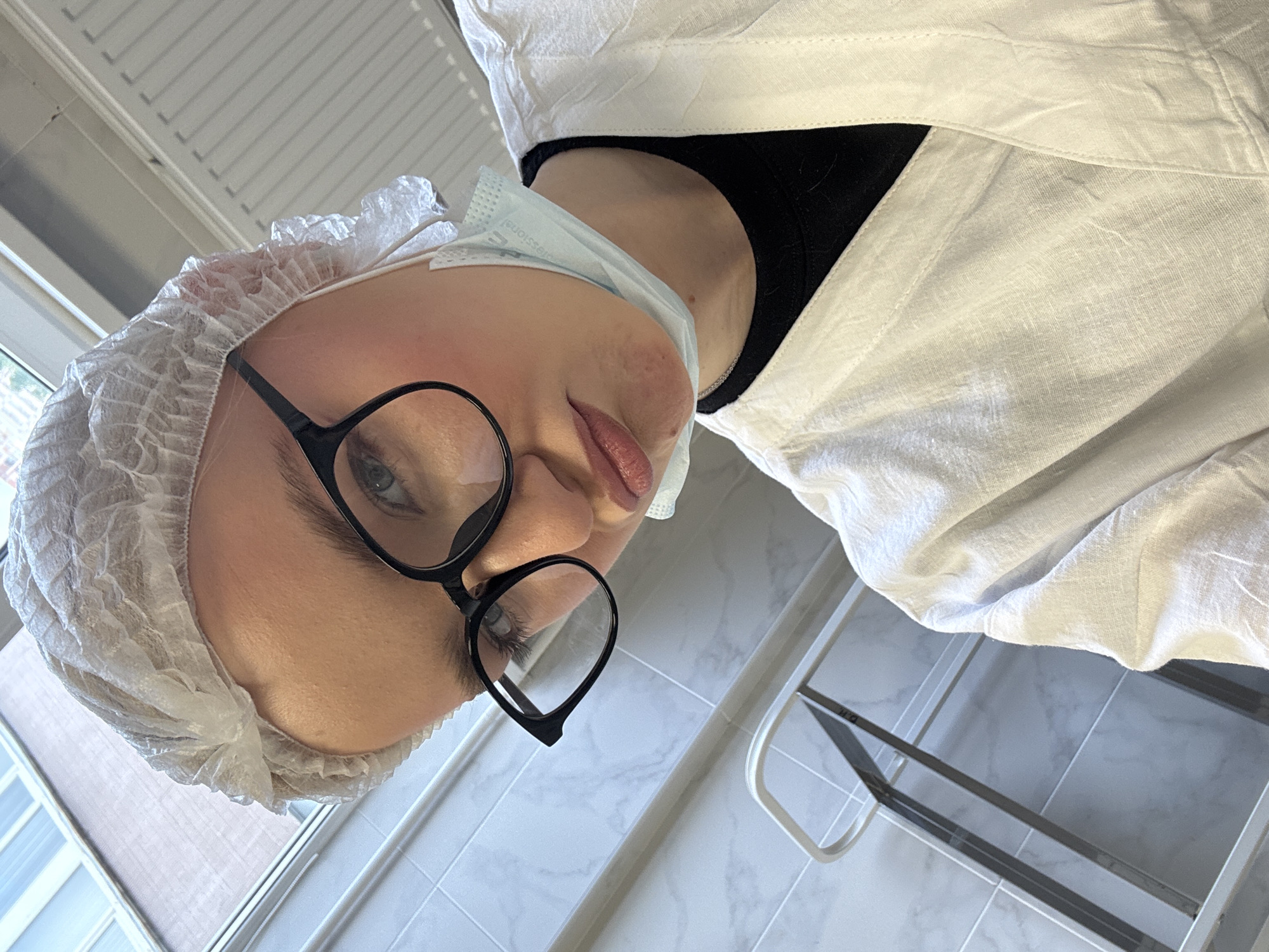 A young woman, wearing a disposable medical hair cap, black-rimmed glasses, and a white medical coat over a black garment, poses for a selfie. A light blue surgical mask is visible, hanging loosely below her chin. She is looking down at the camera with pursed lips. The background appears to be an indoor setting, with tiled walls and what looks like a radiator and a window on the left side. A metal railing or equipment is visible in the lower left corner. The lighting suggests an interior environment, possibly a medical or laboratory setting.