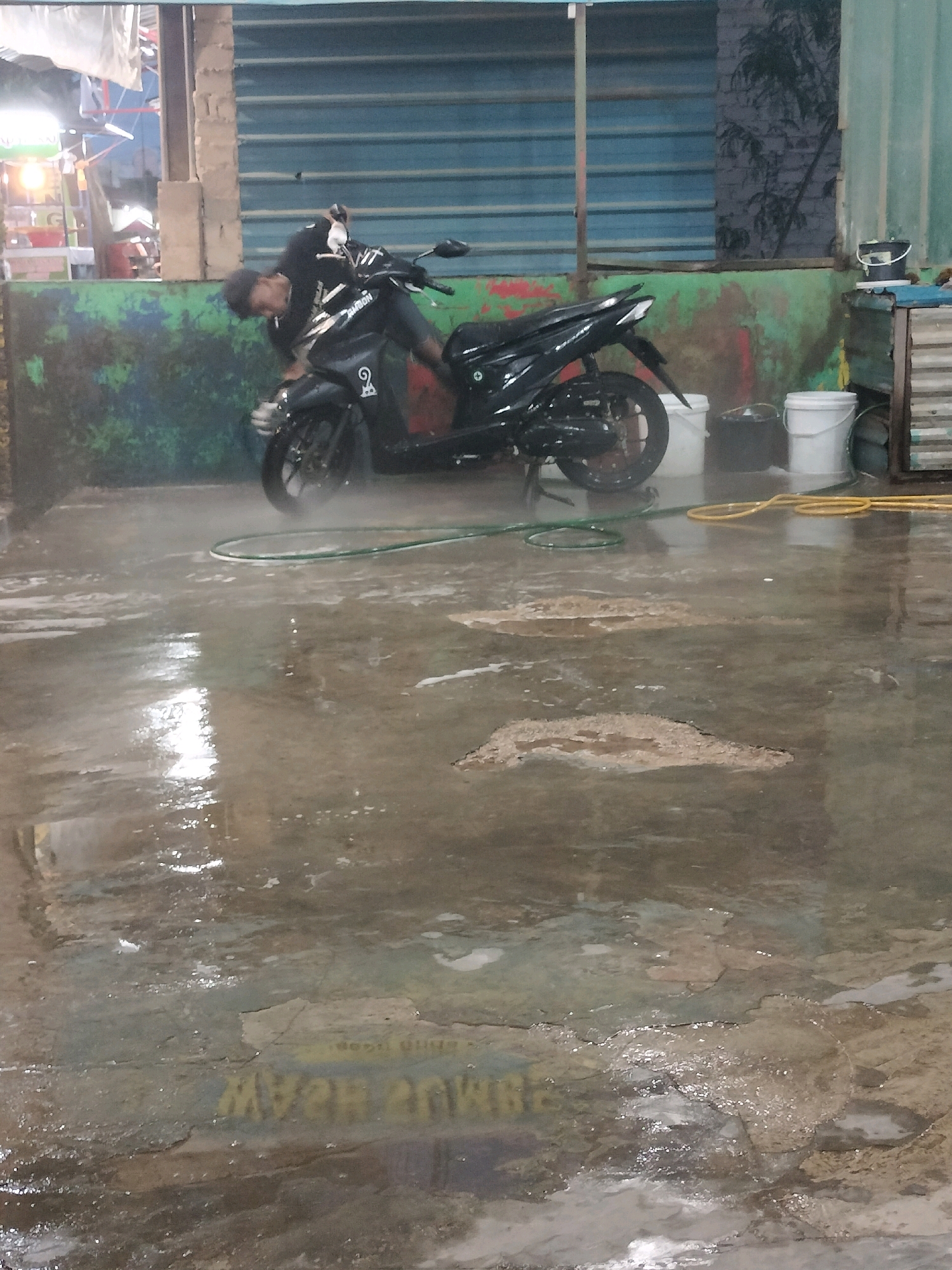 A man is washing a black motorcycle in what appears to be an outdoor or semi-outdoor area, possibly a car wash station, in Batam, Indonesia. The ground is wet and reflective, with puddles of water and some scattered debris. A green hose is visible on the ground, near the motorcycle's front wheel, and a yellow hose is coiled further back. Two buckets, one black and one white, are present behind the motorcycle. The background shows a weathered green wall with some red and white paint splatters. Above the wall is a blue corrugated metal structure. The lighting suggests it might be evening or a dimly lit day. The man, wearing a dark shirt and shorts, is bent over the motorcycle, actively engaged in cleaning it. The motorcycle is a scooter-type, with its kickstand down. The overall impression is a typical scene of motorcycle maintenance in a humid, tropical environment.