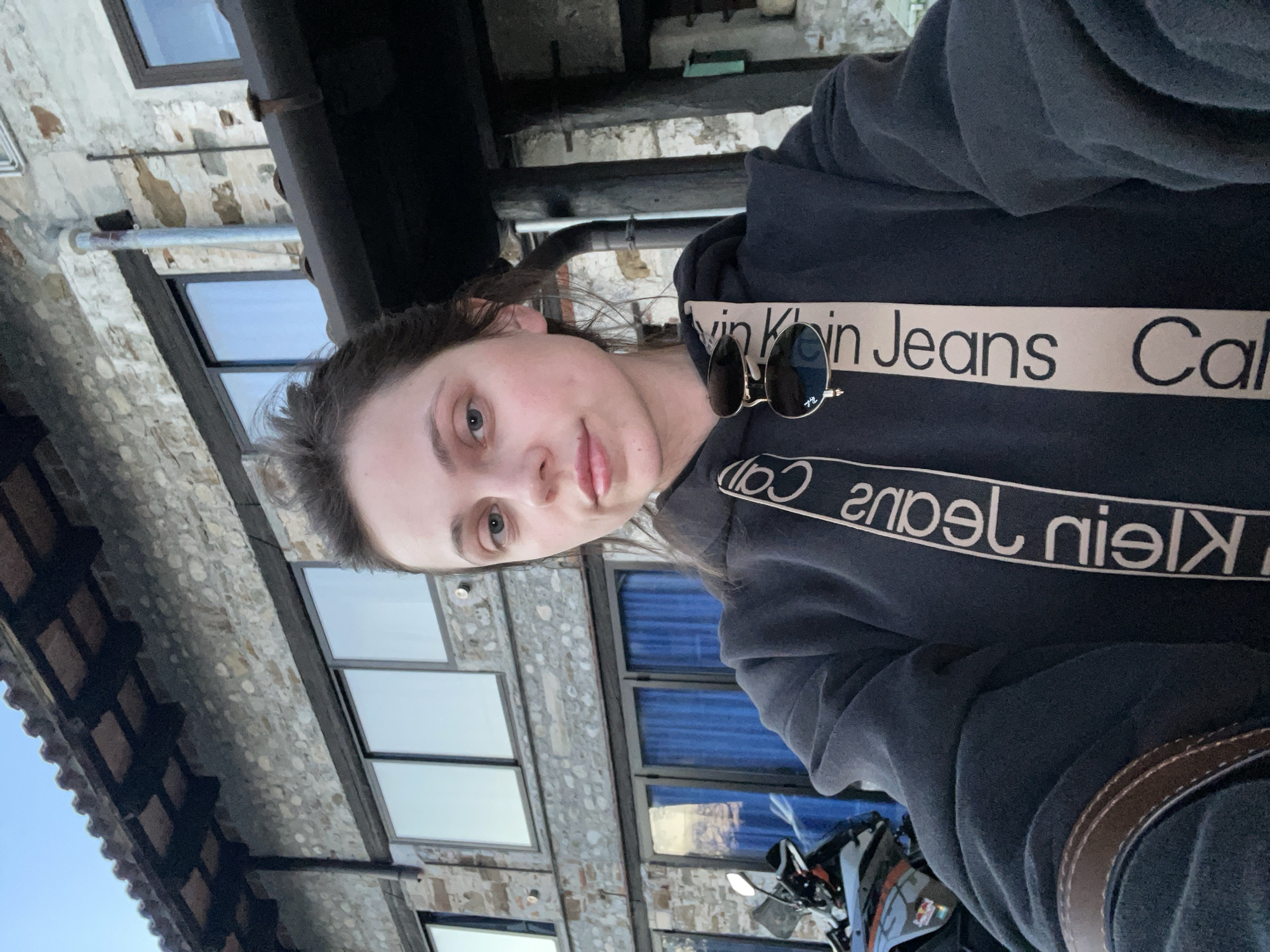 A woman wearing a black Calvin Klein Jeans hoodie poses in front of a weathered stone building with a multi-paned glass window façade. Sunglasses are clipped to the front of the hoodie. A motorcycle is visible in the lower portion of the frame. The image orientation suggests a selfie taken while riding on the motorcycle. The building’s architectural style is consistent with buildings in San Lorenzo, Italy, with its stone construction and window design.