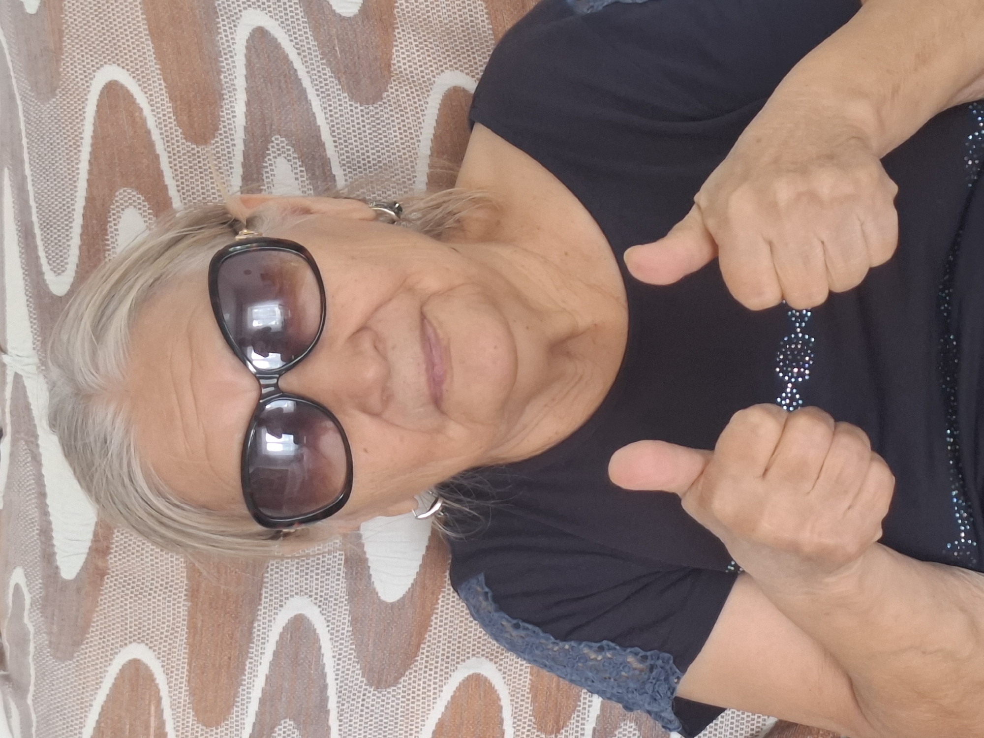 A close-up, waist-up shot shows an elderly woman with grey hair, wearing large black sunglasses, a black shirt with blue lace detailing on the shoulders and a jeweled pattern across the chest, and hoop earrings. She is sitting on a couch with a patterned upholstery of beige and brown wavy lines. The woman is looking directly at the camera with a neutral expression, holding up both thumbs in a "thumbs-up" gesture. The reflection in her sunglasses shows a window and what appears to be a ceiling light. The overall scene is an indoor setting. There is no identifiable location in the city of Almaty, Kazakhstan visible in the image.