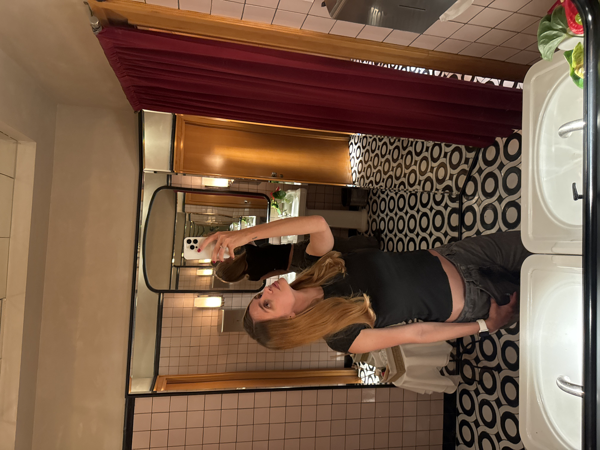 A woman is taking a selfie in a bathroom. The bathroom has pink tile walls, black and white patterned floors, and a red curtain along the top of the mirror and cabinet. There is a double sink on the right of the frame. Without additional information, it is impossible to determine if this location is in Yerevan, Armenia.