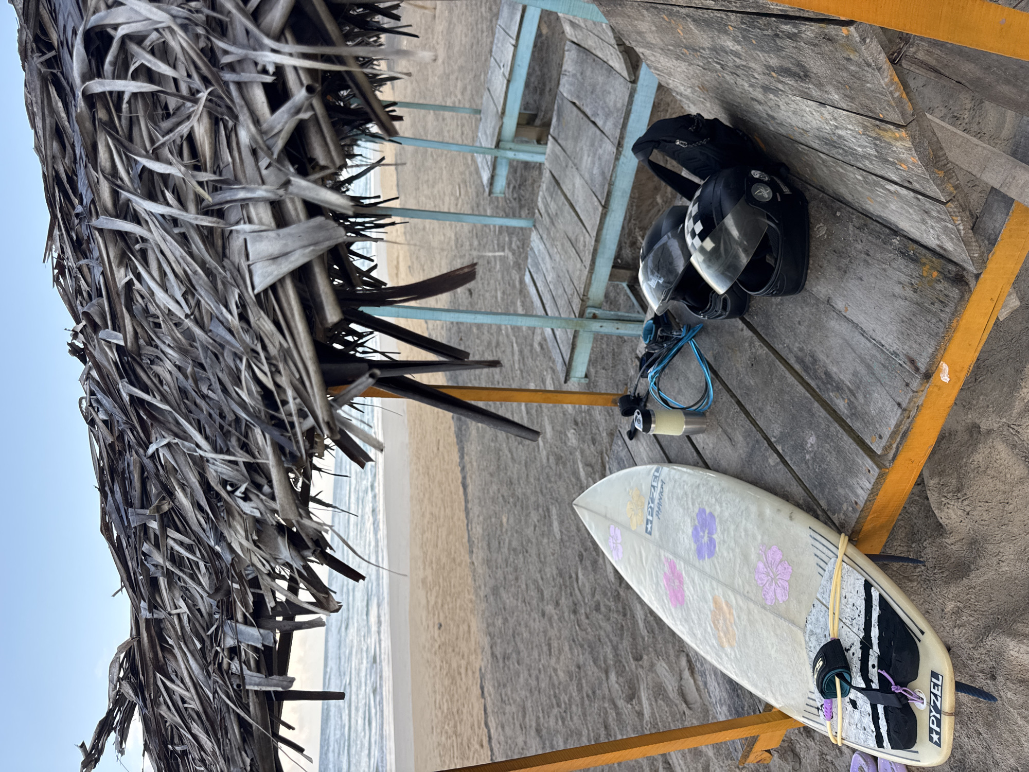 In Kathaluwa, Sri Lanka, a rustic beach scene is depicted under a thatched-roof shelter. A surfboard, adorned with floral patterns, rests on a wooden platform. Nearby, two helmets and a water bottle are placed beside a backpack, suggesting recent activity or preparation for an outing. The wooden structure itself appears weathered, with tables and benches suggesting a communal resting area. The sandy beach stretches towards the ocean, where gentle waves roll onto the shore under a pale sky. The environment is natural and tranquil, hinting at a relaxed atmosphere.