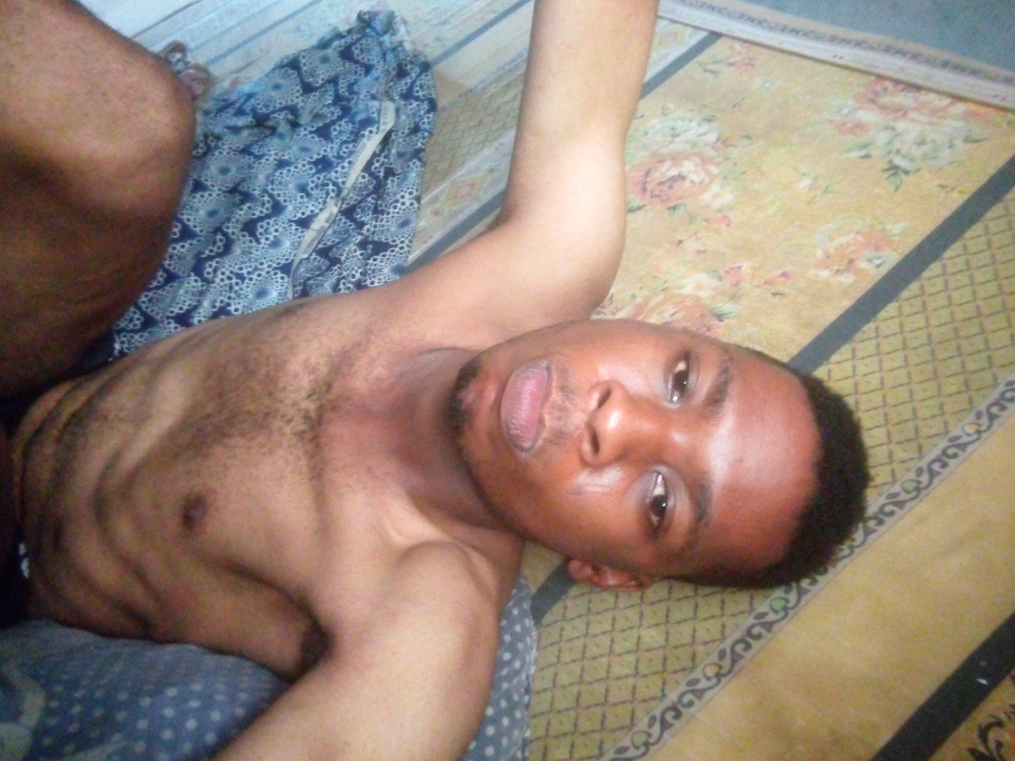 A shirtless person is lying on their back on a bed. The person has dark skin and short, dark hair. Their right arm is raised above their head. The bed has a blue patterned sheet and a yellow and brown floral patterned bedspread. The location is Unknown, Unknown.