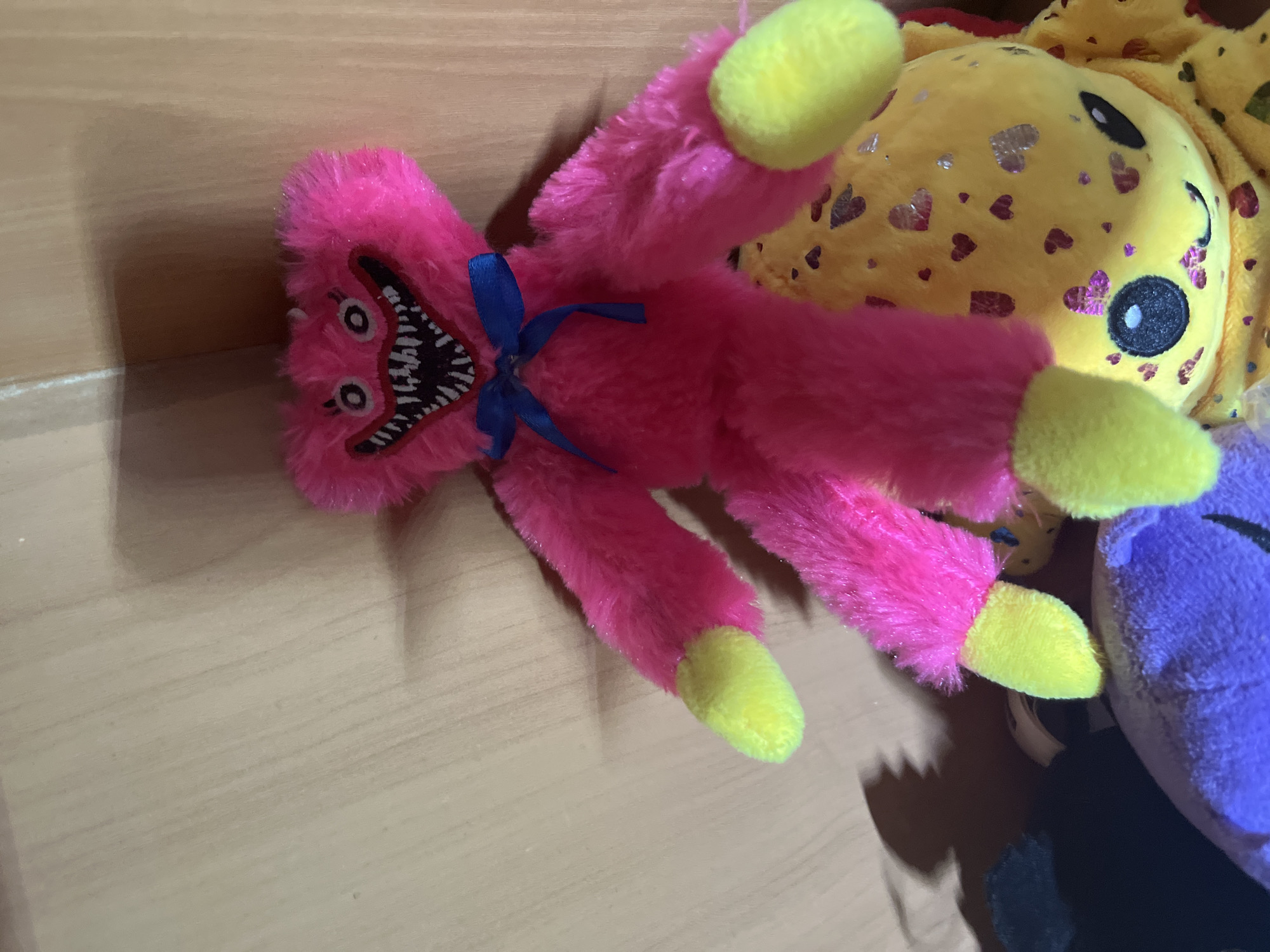 A stuffed toy with bright pink fur and a cartoonishly scary face is positioned on top of a yellow stuffed toy. The pink toy has a blue ribbon tied around its neck and yellow paws. The yellow toy is decorated with glittery heart shapes and has large black and white eyes. The background appears to be a light-colored wooden surface, possibly a shelf or wall. The lighting suggests an indoor environment. No specific location within Ashkadar, Russia, can be determined from the image.