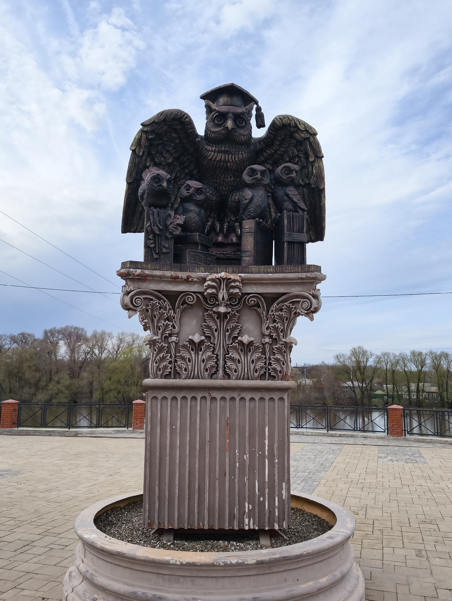 The image shows a statue in the city of Balashov, Russia. The statue is of an owl wearing a graduation cap, surrounded by smaller owls, books, and a chair. The statue is placed atop a classical-style column, which sits in a circular base filled with gravel. The column is decorated with acanthus leaves and other classical motifs. The statue is located on a paved plaza with a river and trees in the background. A fence and some electrical wires are also visible. The sky is cloudy.