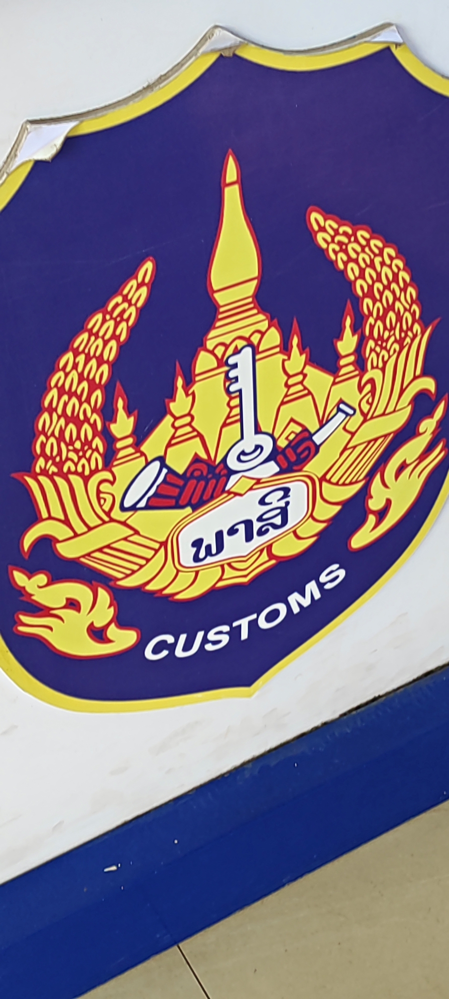 This image shows a close-up of a logo on a surface. The logo is shield-shaped with a purple background and a yellow border. It features a central design in yellow and red depicting a temple-like structure, a key, and what appears to be ears of rice or wheat. Below the central design, there are two lines of text. The top line is in a script that appears to be Lao, and the bottom line reads "CUSTOMS" in white English letters. The logo shows some wear and tear, with peeling edges. The background outside the logo is a white surface with a blue strip along the bottom edge. The image is slightly angled and vertically oriented. There are no people or discernible environmental details indicating a specific location within the city of Xayden, Laos. The image focuses solely on the logo and the surface it is affixed to.