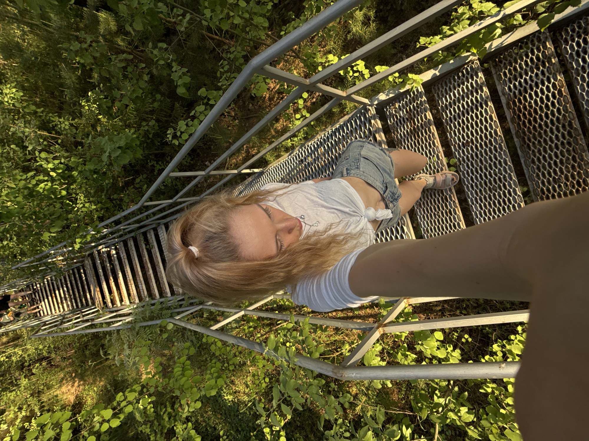 A high-angle, close-up shot depicts a woman standing on metal stairs with grated steps, seemingly part of a tower or tall structure. The woman, wearing denim shorts and a white top, is positioned towards the right side of the frame. Her blonde hair is pulled back into a ponytail. She is holding the camera to take a selfie. The stairs descend steeply, and through the grating, dense green foliage is visible. The surrounding environment suggests a wooded area. The location is in the city of Noviy, Russia. In the distance a few people can be seen walking on the stairs.
