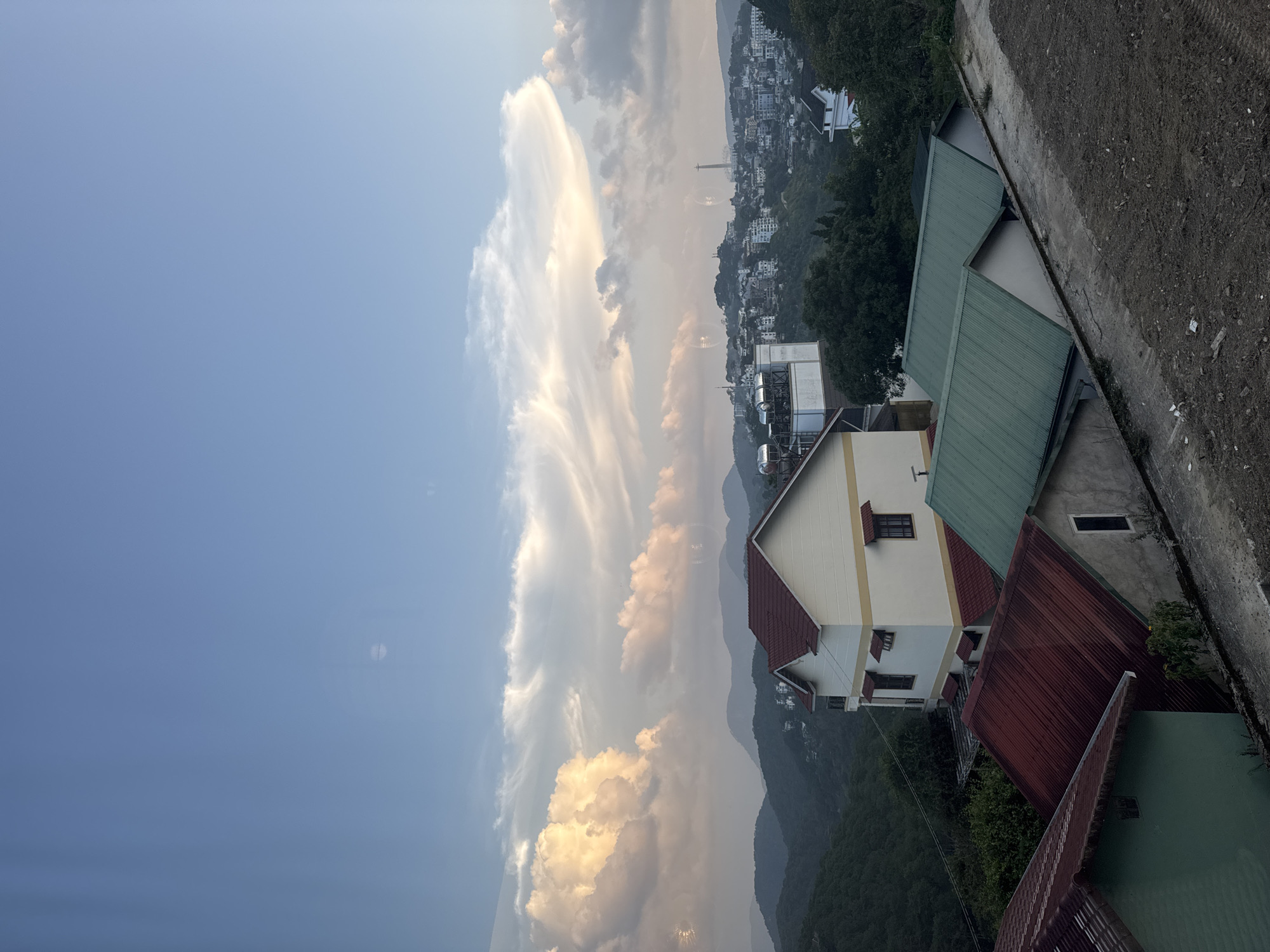 The image captures a hillside view of Da Lat, Vietnam, characterized by its characteristic rolling terrain and a mix of residential structures. In the foreground, several houses with distinct roofing styles are visible. A house with a prominent cream-colored facade and a red-tiled roof stands out, featuring multiple windows and awnings. Adjacent to it are other houses, one with a red corrugated metal roof and another with a green corrugated metal roof, suggesting a varied architectural landscape.
The mid-ground reveals a denser urban area cascading down the hills, dotted with numerous buildings of various sizes and colors, predominantly white and light-colored. A tall industrial structure, possibly a communication tower or a chimney, rises above the city in the distance, a common sight in many developing urban centers. The background is dominated by layered mountain ranges, softened by the hazy atmosphere characteristic of Vietnamese highlands.
The sky above is a spectacle of dramatic cloud formations. Cumulus clouds, illuminated by what appears to be late afternoon or early morning sunlight, are tinged with warm oranges and yellows, contrasting with the cooler blue of the clearer sky. Wispy cirrus clouds stretch across the expanse, adding texture and depth to the sky. The overall impression is one of a serene, yet busy, urban environment nestled within a majestic natural landscape. There are no people visible in the image. The scene appears to be captured from a slightly elevated vantage point, possibly a balcony or a higher floor of a building, overlooking the town and the surrounding mountains.