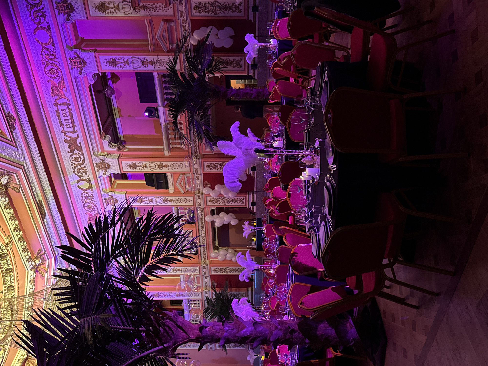 The image captures an elegantly set indoor venue, likely a grand hall or ballroom, prepared for a formal event such as a dinner or reception. A long banquet table, adorned with place settings including plates, wine glasses, and cutlery, extends diagonally across the scene. The table is decorated with tall white feather centerpieces and smaller candles, illuminated by the ambient lighting. Reddish-pink upholstered chairs with golden frames are arranged along the table.
Large, dark palm trees are strategically placed throughout the room, their fronds reaching upwards, adding a tropical or exotic touch to the decor. In the background, clusters of white balloons resemble clouds, contributing to a festive atmosphere.
The architecture of the venue is highly ornate, featuring elaborate moldings, decorative columns, and gold accents on the walls and ceiling, painted in shades of pink and orange. The space is dramatically lit with vibrant purple and pink artificial lighting, creating a luxurious and celebratory mood. Spotlights are visible on the upper walls.
On a prominent wall panel, the inscription "13 III 1913" is visible, likely denoting a significant date for the building or event. A person is partially visible in the background, near one of the palm trees. The overall impression is that of an upscale evening event, possibly taking place in a historic and ornate venue in Sofia, Bulgaria.