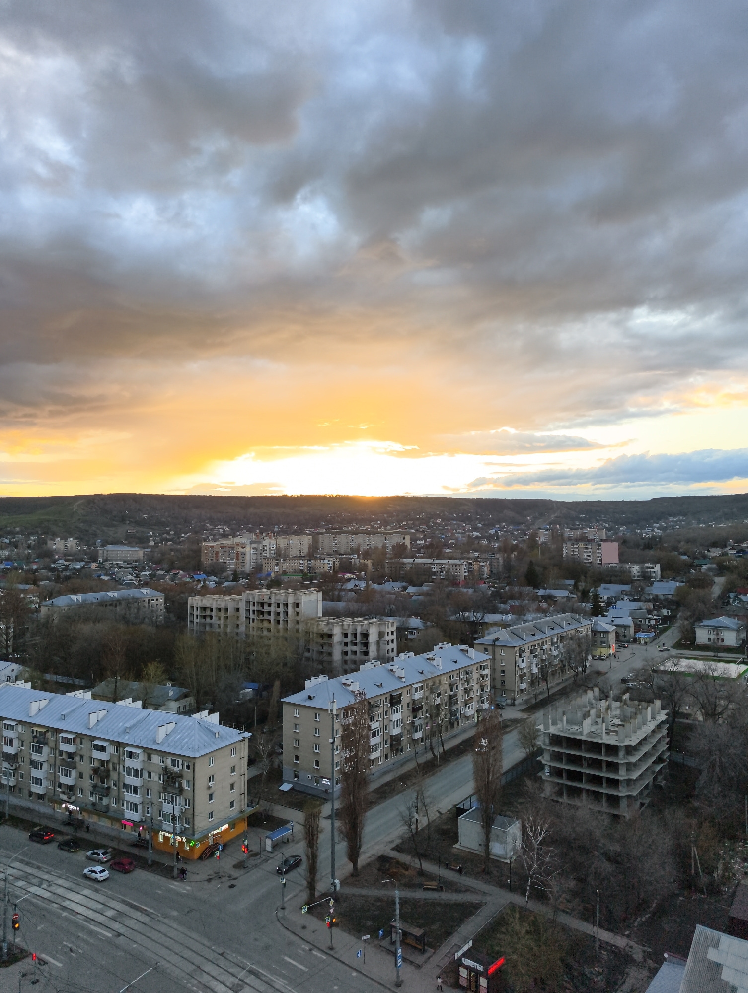 The image shows an urban landscape in Saratov, Russia, under a cloudy sky with a bright sunset or sunrise on the horizon. In the foreground are residential buildings, typically Soviet-era apartment blocks, with gray facades and balconies. A street intersection is visible with pedestrian crossings, traffic lights, and tram tracks. There are a few cars on the road and people walking near a corner pharmacy ("Apteka"). An unfinished concrete building structure sits to the right, indicating ongoing construction in the area. The overall scene conveys a sense of a city waking up or settling down for the evening.