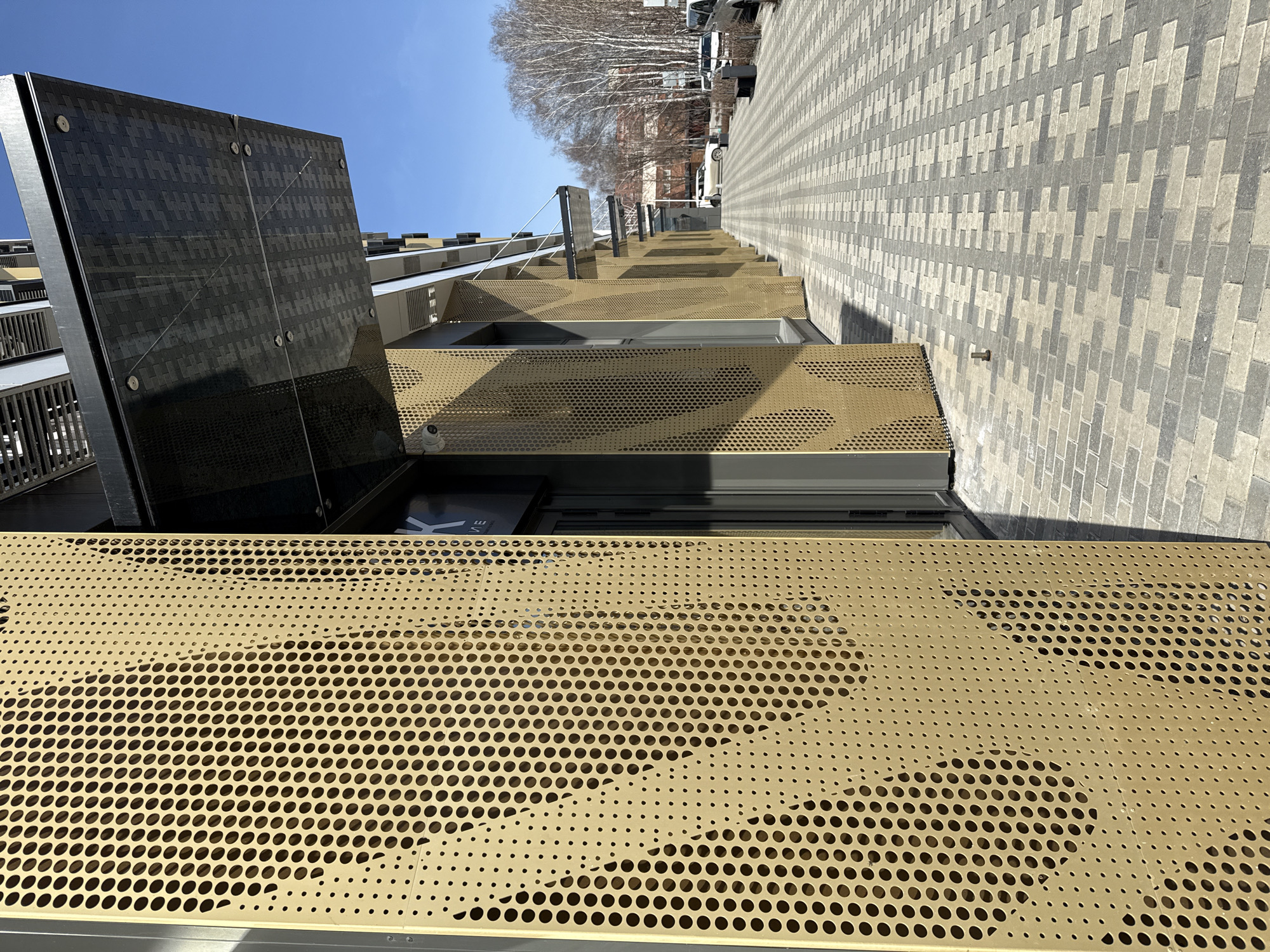 The image depicts a modern building facade in what appears to be a residential area. The location is identified as Yekaterinburg, Russia. The building features a multi-layered design, with dark paneled sections and perforated golden screens with varying hole density. The facade angles away, revealing a pedestrian walkway composed of patterned gray and white pavers leading towards trees and parked vehicles in the distance. The scene is illuminated by bright sunlight under a clear blue sky.