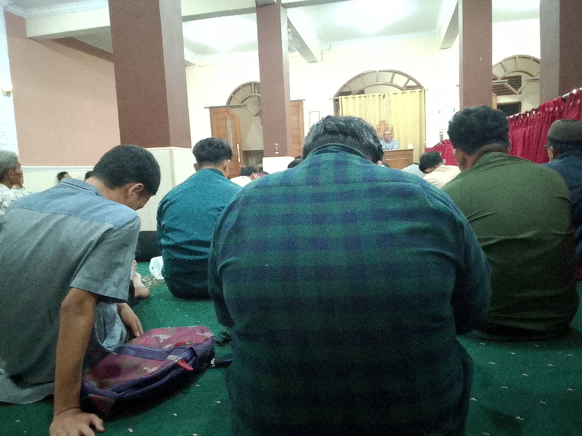 In Kertajaya, Indonesia, a group of men are seated on a green carpet in what appears to be a prayer hall. The room is large with high ceilings supported by brown columns. In the background, a man is speaking from a raised platform in front of an arched doorway draped with yellow curtains. The men in the foreground are facing away from the camera, engrossed in the prayer or sermon. Some are wearing traditional Islamic attire, while others are in casual shirts. Bags and other personal items are scattered around them on the carpet. The lighting in the room is soft, suggesting it could be daytime or an indoor event.