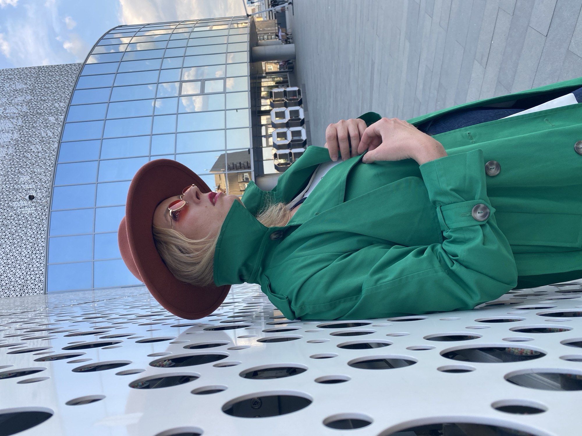 A person wearing a brown hat and a green trench coat is lying on a white surface with circular cutouts. The person also wears small, round, tinted glasses. The background shows modern architecture with reflective glass panels and a sign with the number "8881". The scene is in Yekaterinburg, Russia, as indicated by the architectural style and signage.