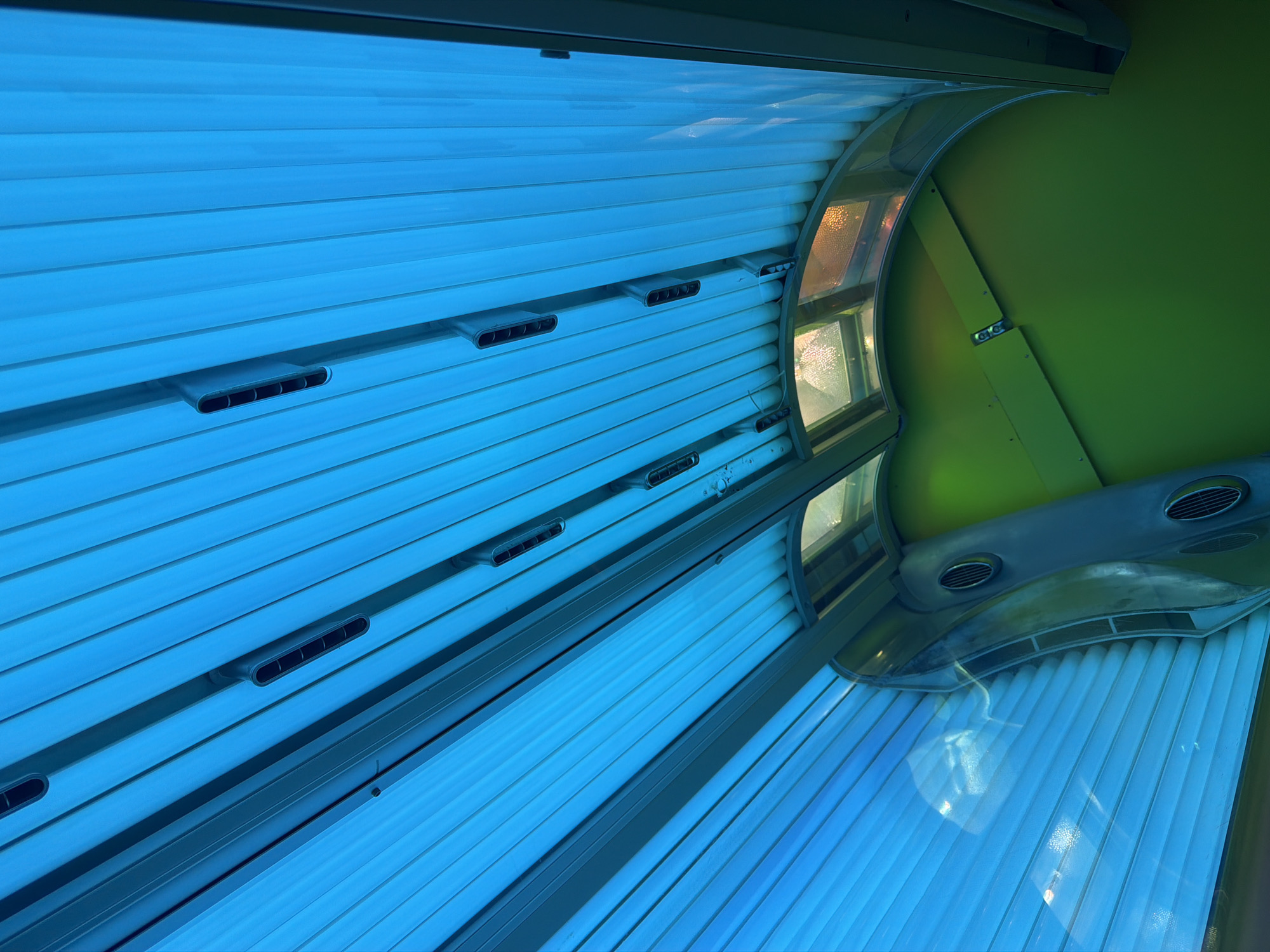 The image shows the interior of a tanning bed. The bed is illuminated with bright blue UV lights, and a green surface is visible in the background. Several air vents are embedded in the tanning bed's structure. There are no people or any indication of the location in Kyiv, Ukraine.