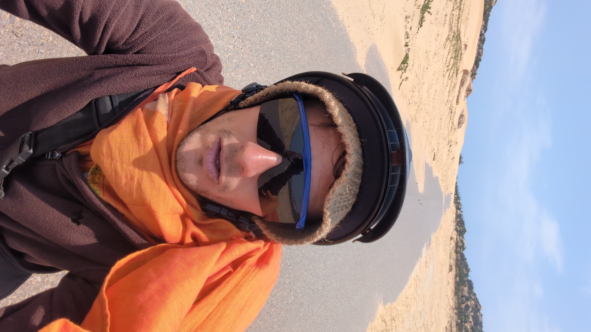A selfie taken in Phuoc Dinh, Vietnam, shows a person wearing a brown fleece jacket, an orange scarf, a black helmet, and blue-framed sunglasses. The background consists of a sandy area, possibly dunes, meeting a clear blue sky. The person is likely standing on a paved surface.