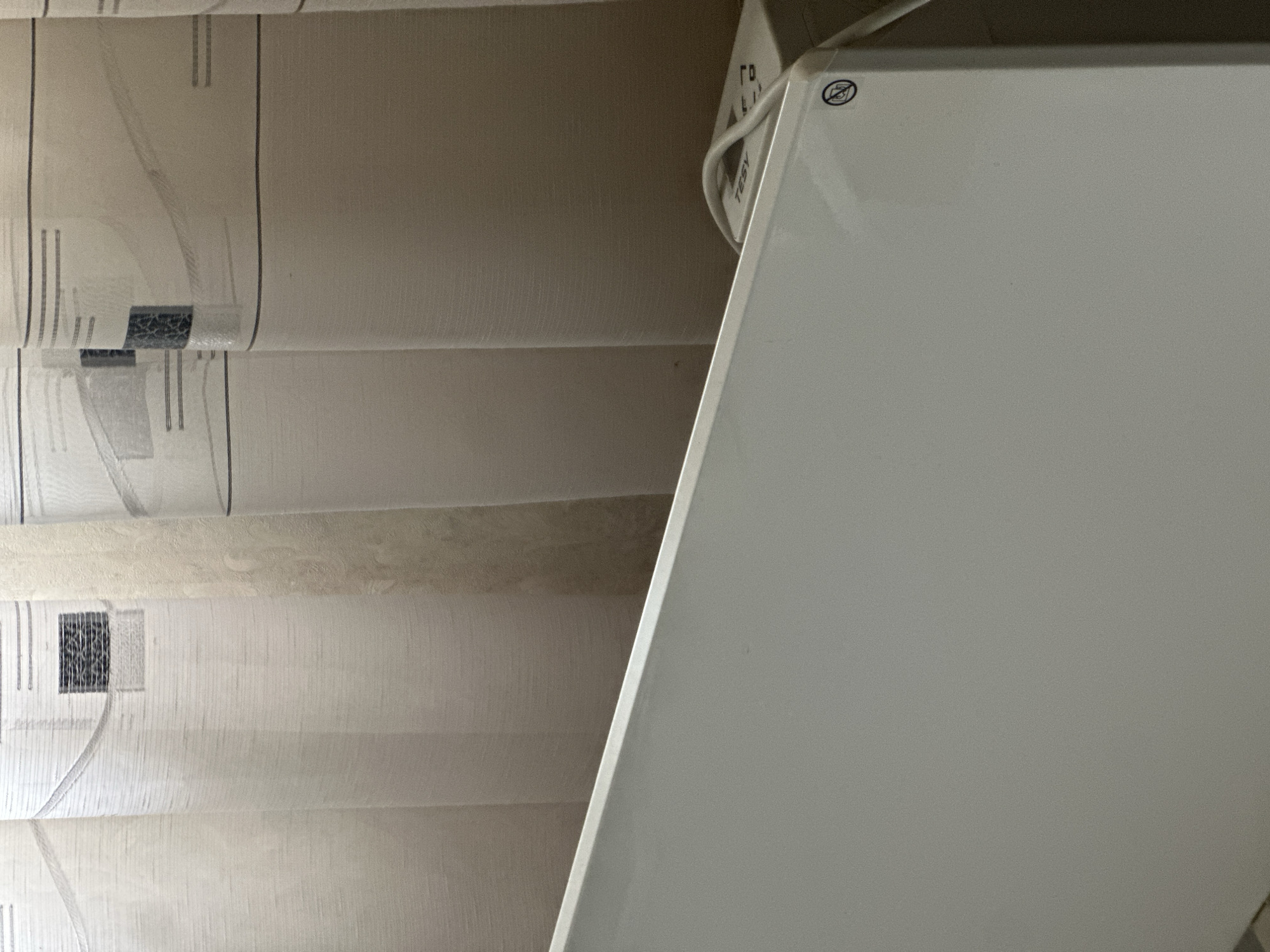 The image presents a close-up view of a white TESY panel heater positioned adjacent to a beige Roman blind. The blind features a repeating pattern of vertical lines and geometric shapes. The heater occupies the right side of the frame, while the blind is on the left. The setting appears to be indoors. No specific details indicate the location is in Slavyani, Bulgaria.