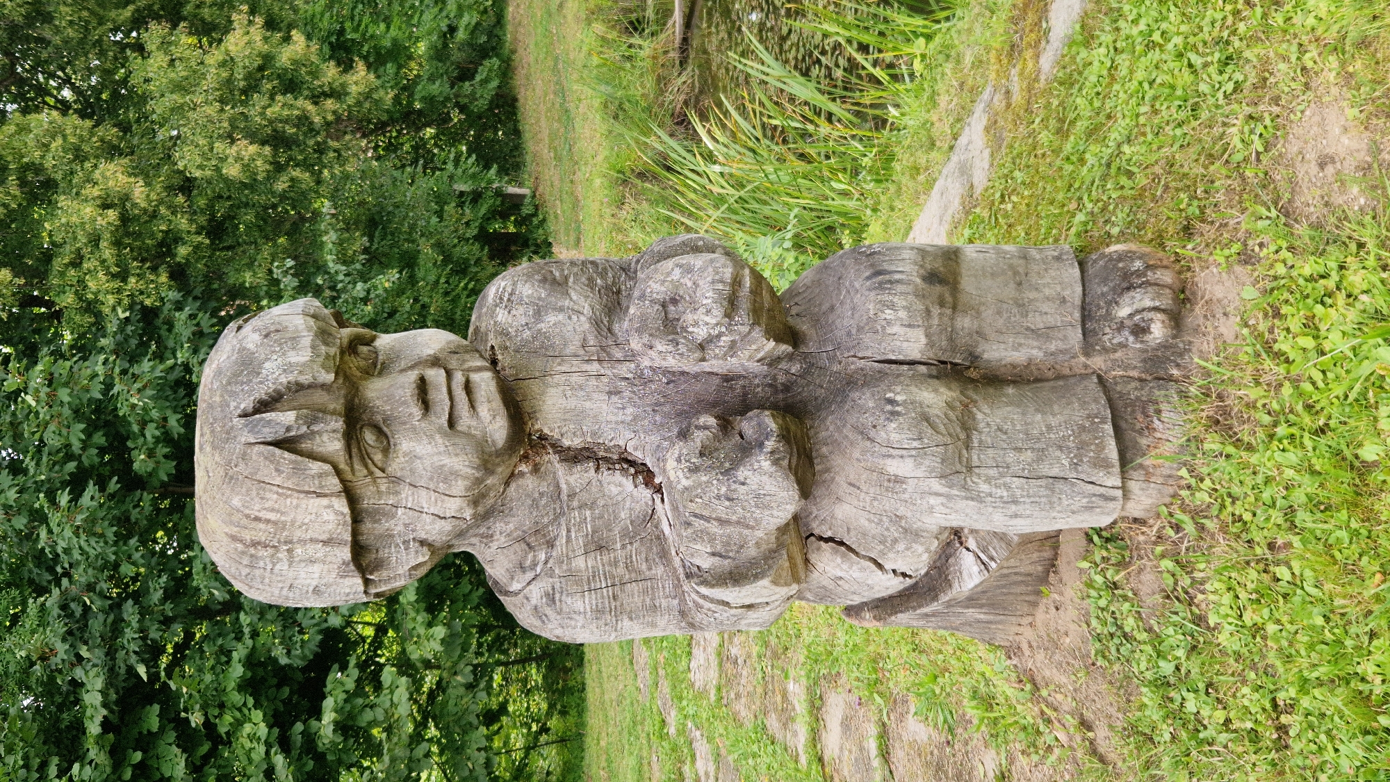 A weathered wooden sculpture of a child sits in a grassy area with trees in the background. The sculpture is of a seated figure with a bowl-shaped haircut, large hands, and detailed legs and feet. The wood shows significant cracking and signs of age. To the right of the sculpture, there is a body of water with reeds growing along its edge. A stone path is visible on the left and in the background. The sculpture is located in Kazdanga, Latvia, specifically in the park adjacent to Kazdanga Manor.