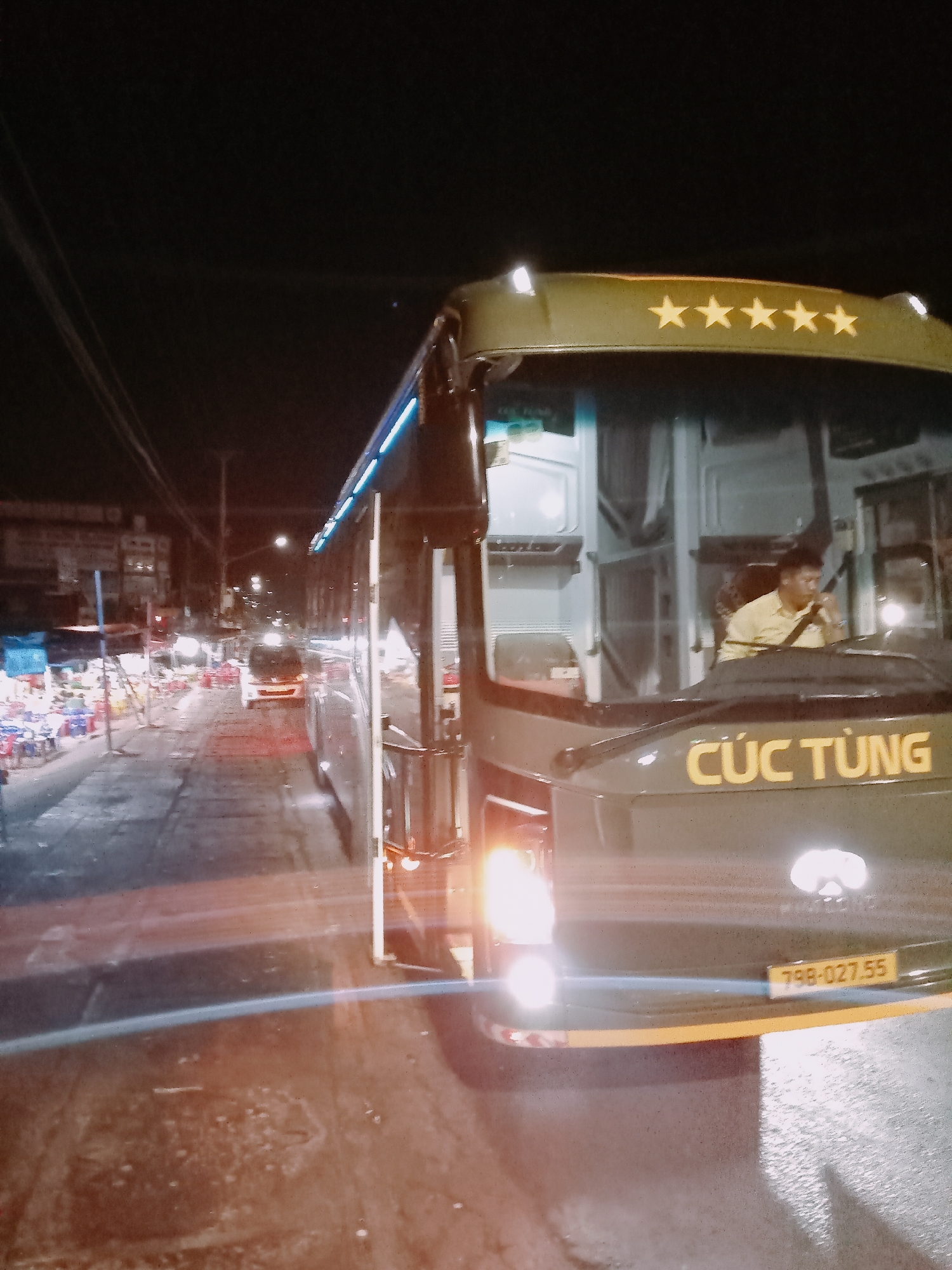 A nighttime shot depicts a Cúc Tùng bus, its headlights on, positioned on a street in Ea Drăng, Vietnam. The bus, dark green with the name "Cúc Tùng" in yellow on its front, also features five stars above the windshield. The license plate reads "79B-027.55". A man in a light-colored shirt is seen driving the bus. The street is wet and reflects light. On the left, tables and chairs line the sidewalk, suggesting a restaurant or gathering place. In the distance, a few vehicles and streetlights illuminate the street.
