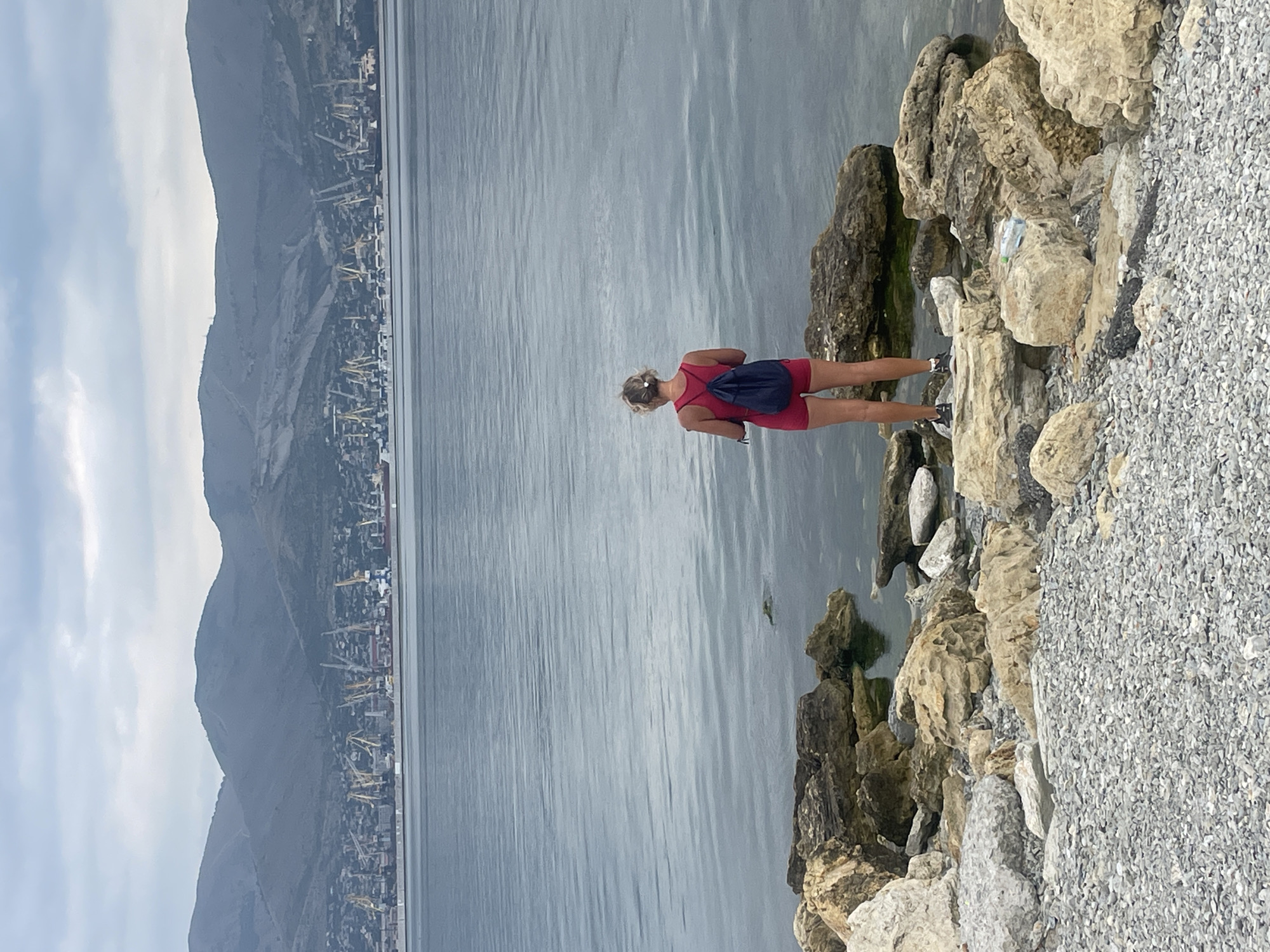 A woman with blonde hair, seen from behind, stands on a rocky shore facing a calm body of water. She is wearing a red tank top and red shorts, with a dark blue backpack slung over her shoulders. Her dark sneakers are visible. The shore is covered in small grey pebbles and scattered large rocks. The water extends to a distant industrial port with numerous cranes and ships lining the coast. Behind the port, rolling, barren hills rise under an overcast sky. The scene appears to be the Novorossiysk bay in Russia, likely near the commercial port.
