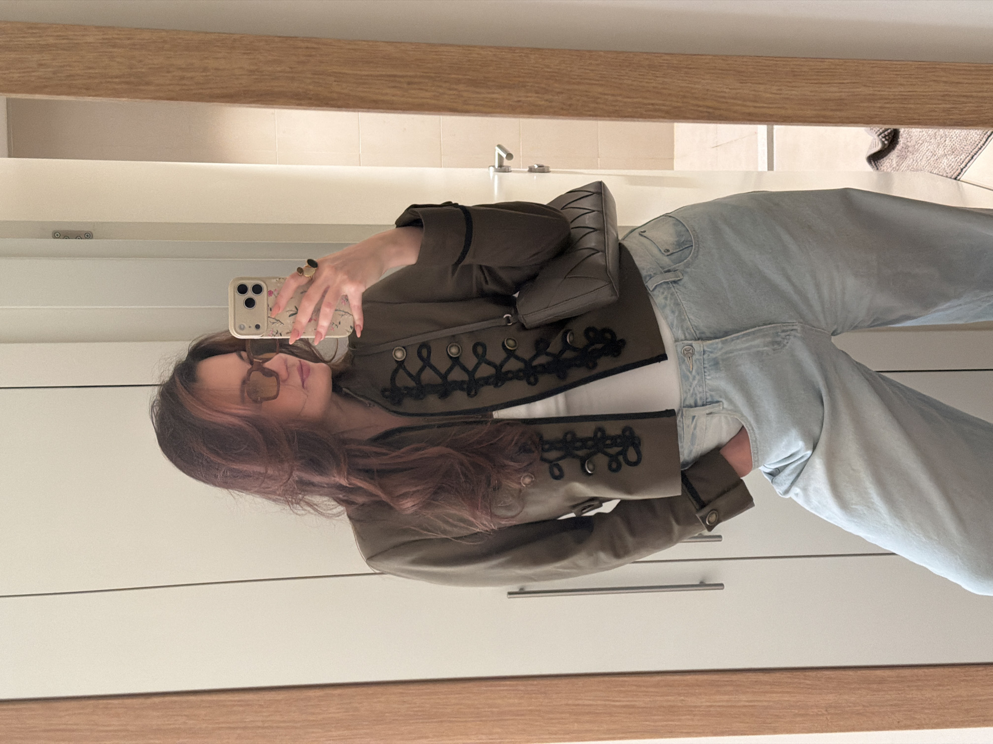 A young woman takes a selfie in a bathroom mirror. She wears a khaki jacket with dark embellishments and light blue jeans. She holds a smartphone with a decorative case. The bathroom features white cabinets with simple handles and a light wood-framed mirror. The environment is well-lit and suggests a modern interior. The city of Belgrade, Serbia, cannot be confirmed based on visual information.