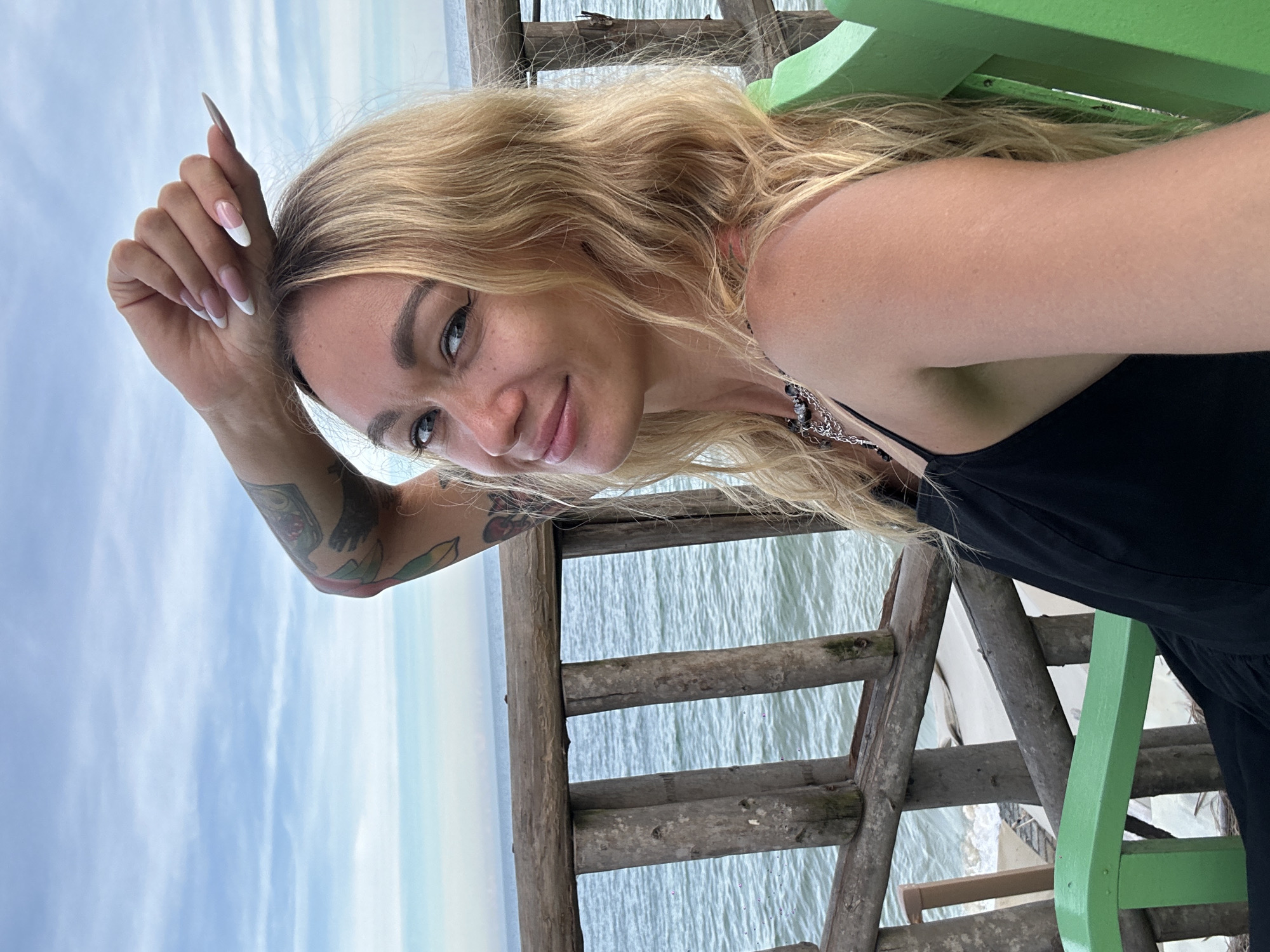 A blonde woman with tattoos on her arm sits on a green chair near the ocean, leaning her arm on her head and looking at the camera. She is wearing a black dress and a necklace. The ocean and a wooden railing are visible behind her. The scene appears to be outdoors, possibly at a beachside resort.