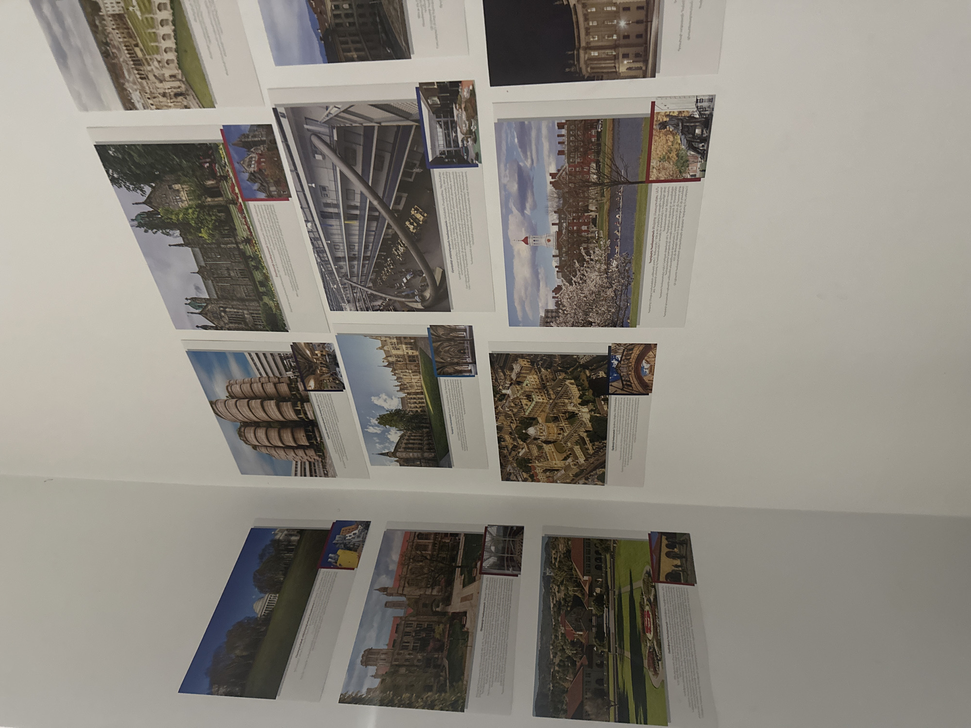 The image shows a section of a white wall displaying numerous framed photographs and accompanying text. The photographs appear to be of architectural structures, possibly university campuses or buildings. The frames are arranged in a grid-like pattern, though not perfectly aligned. The environment is indoors, indicated by the plain white wall, and the lighting is ambient, suggesting the photographs are part of an exhibit or display. There are no people visible in the scene. The content of the photographs suggests a theme of architecture, education, or urban landscapes. No information can be extracted from the image which can give details about Yerevan, Armenia.
